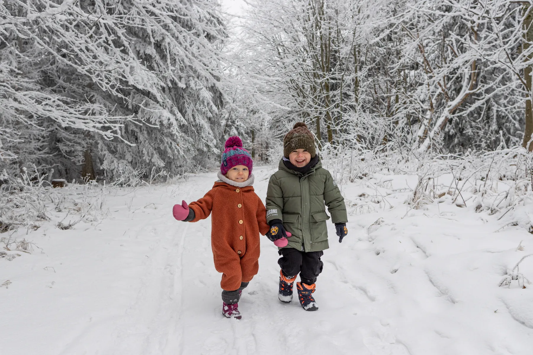 Zwei kleine Kinder gehen auf einem verschneiten Waldweg. Das eine trägt einen braunen Schneeanzug und eine bunte Mütze, das andere einen grünen Mantel und einen braune Mütze. Kahle, verschneite Bäume umgeben sie. Beide lachen in die Kamera.