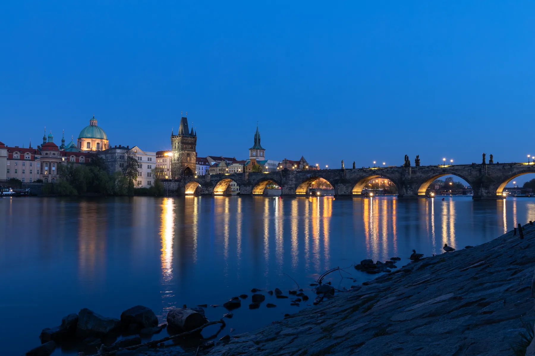 Die Karlsbrücke Prag in abendlicher Beleuchtung zur blauen Stunde und spiegelt sich in der ruhigen Moldau, während die historischen Gebäude und Türme unter einem klaren blauen Himmel zu sehen sind.