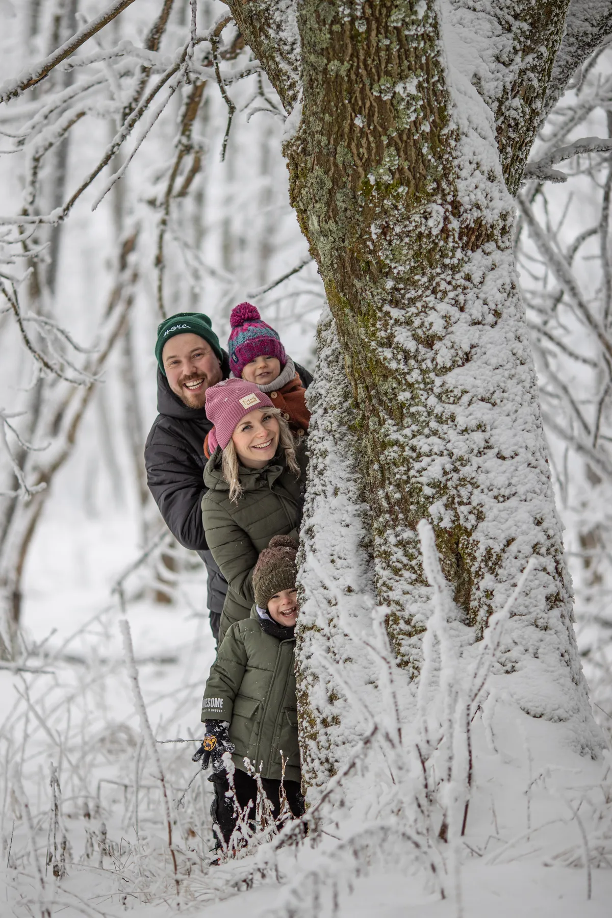 Vier Personen, zwei Erwachsene und zwei Kinder, stehen in Winterkleidung im Freien und schauen hinter einem schneebedeckten Baum in einem verschneiten Wald hervor. Der Schnee bedeckt den Boden und die Äste, und die Menschen lächeln.