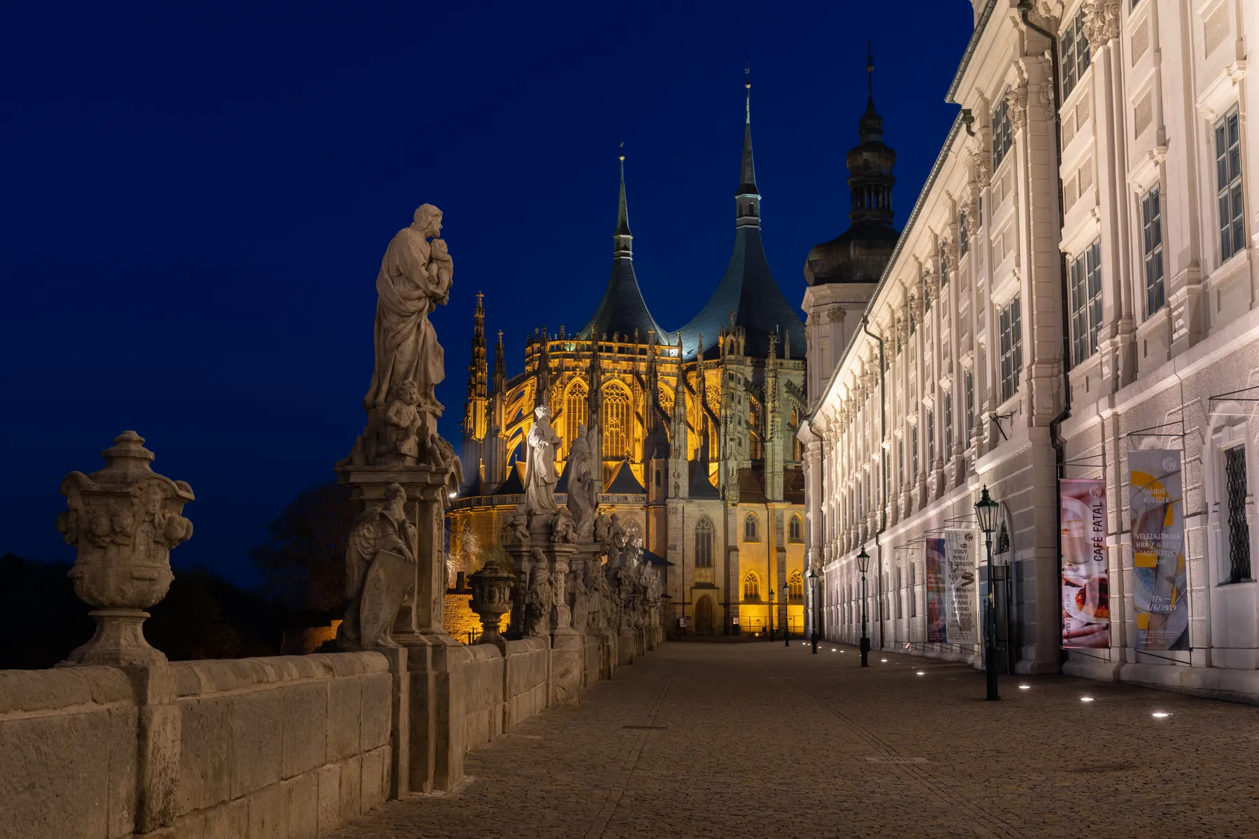 Die hell erleuchtete St. Barbara Kathedrale mit hohen Türmen steht neben einem weißen Barockgebäude in Kutná Hora in Tschechien bei Nacht, von einem mit Statuen und Steingeländern gesäumten Kopfsteinpflasterweg aus betrachtet. Der Nachtimmel ist tiefblau.
