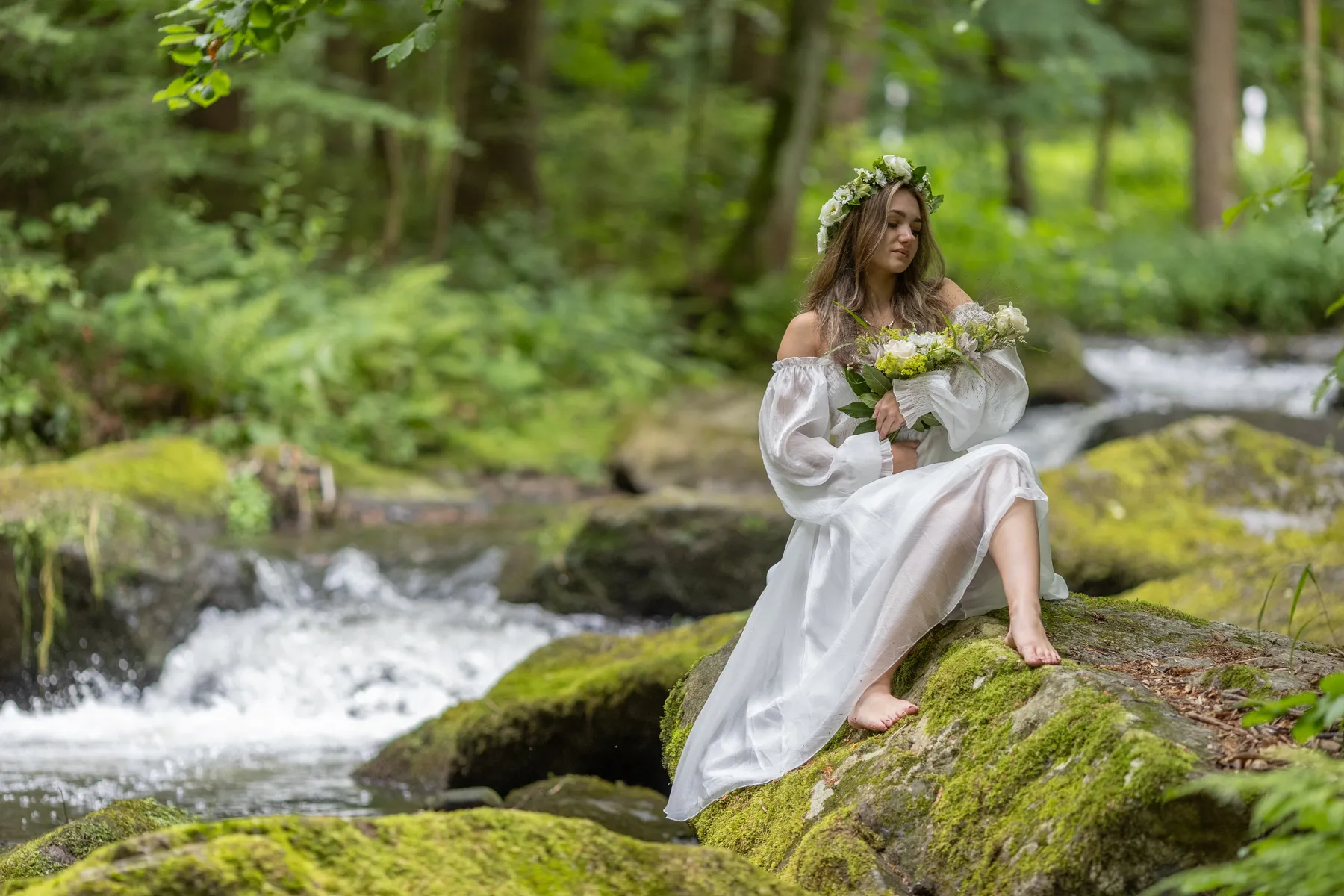 Eine Frau in weißem Kleid und Blumenkranz sitzt barfuß auf einem moosbewachsenen Felsen neben einem fließenden Bach in einem Wald, hält einen Blumenstrauß in der Hand und schaut auf den Blumenstrauß in ihren Armen