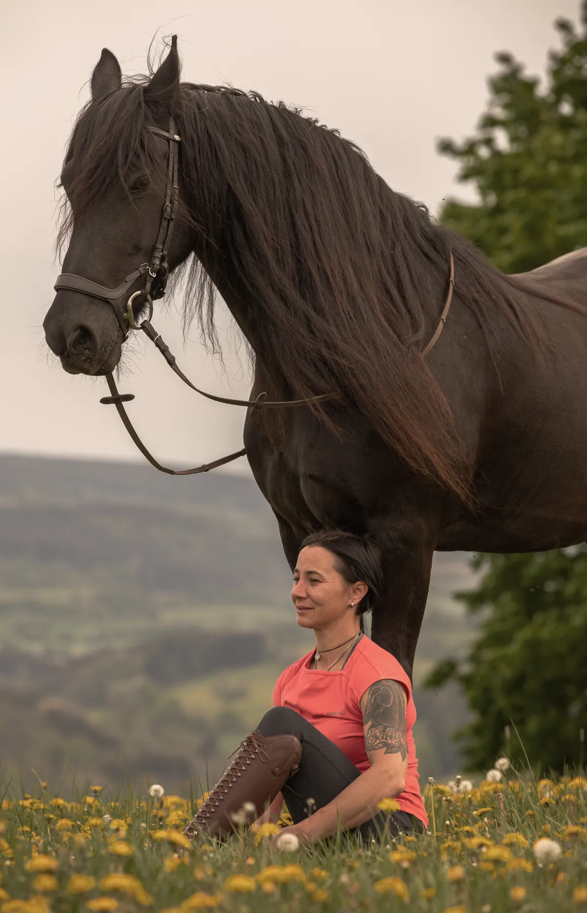 Eine Frau mit kurzen dunklen Haaren und einer Tätowierung auf dem Arm sitzt im Schneidersitz in einem gelben Blumenfeld, während hinter ihr ein großes schwarzes Pferd mit dem Kopf in der Nähe ihres Kopfes vor einer Kulisse aus Hügeln und Bäumen steht.