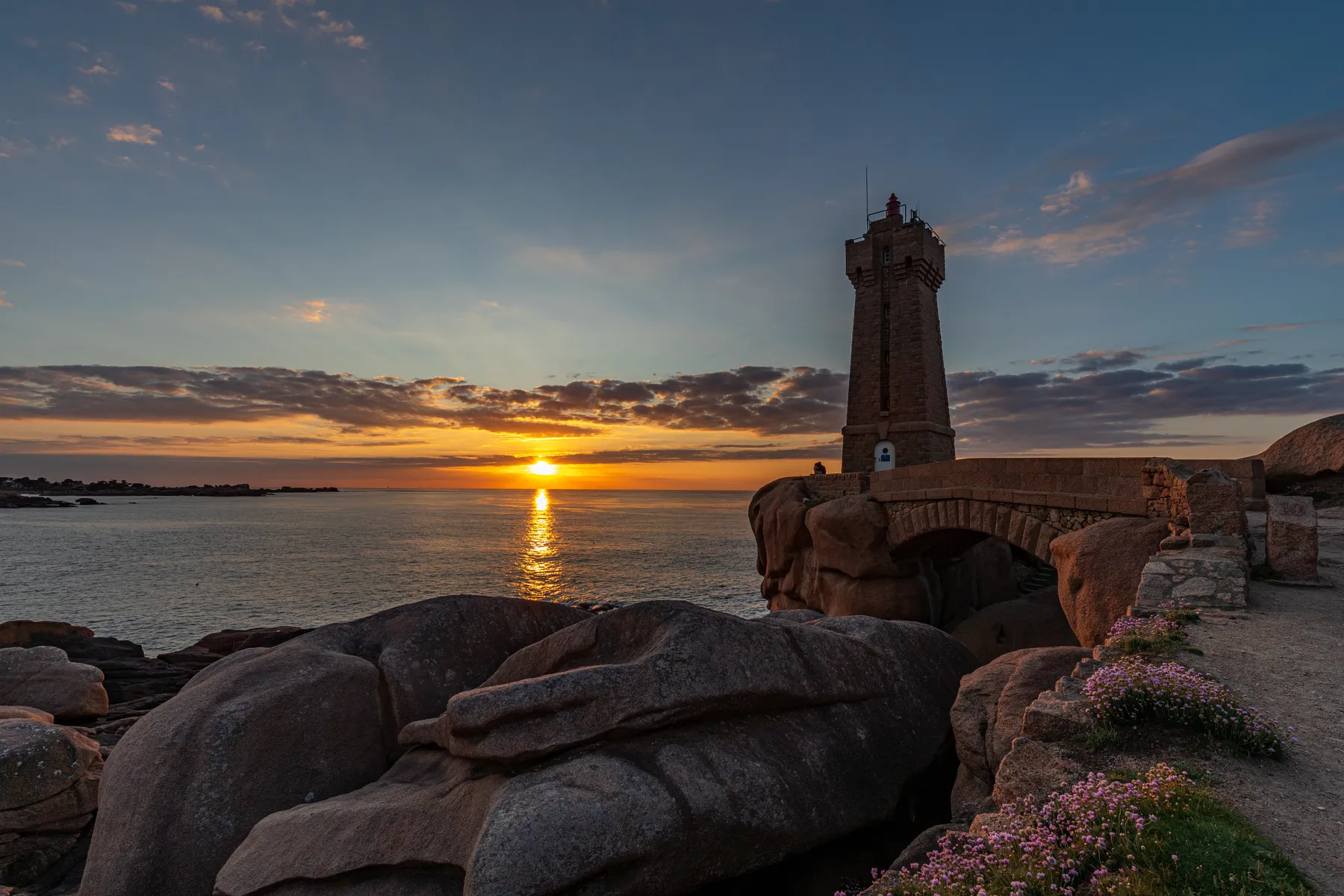 Ein Leuchtturm an der rosa Granitküste der Bretagne. vor Ihm sehen wir eine Brücke aus Stein und riesige Felsblöcke. Das Meer ist ruhig und im Hintergrund versinkt die Sonne und färbt den Horizont gelb. Ein paar Wolken sind am Horizont zu sehen und in der Ferne Silhouetten von Felsen die ins Meer ragen.