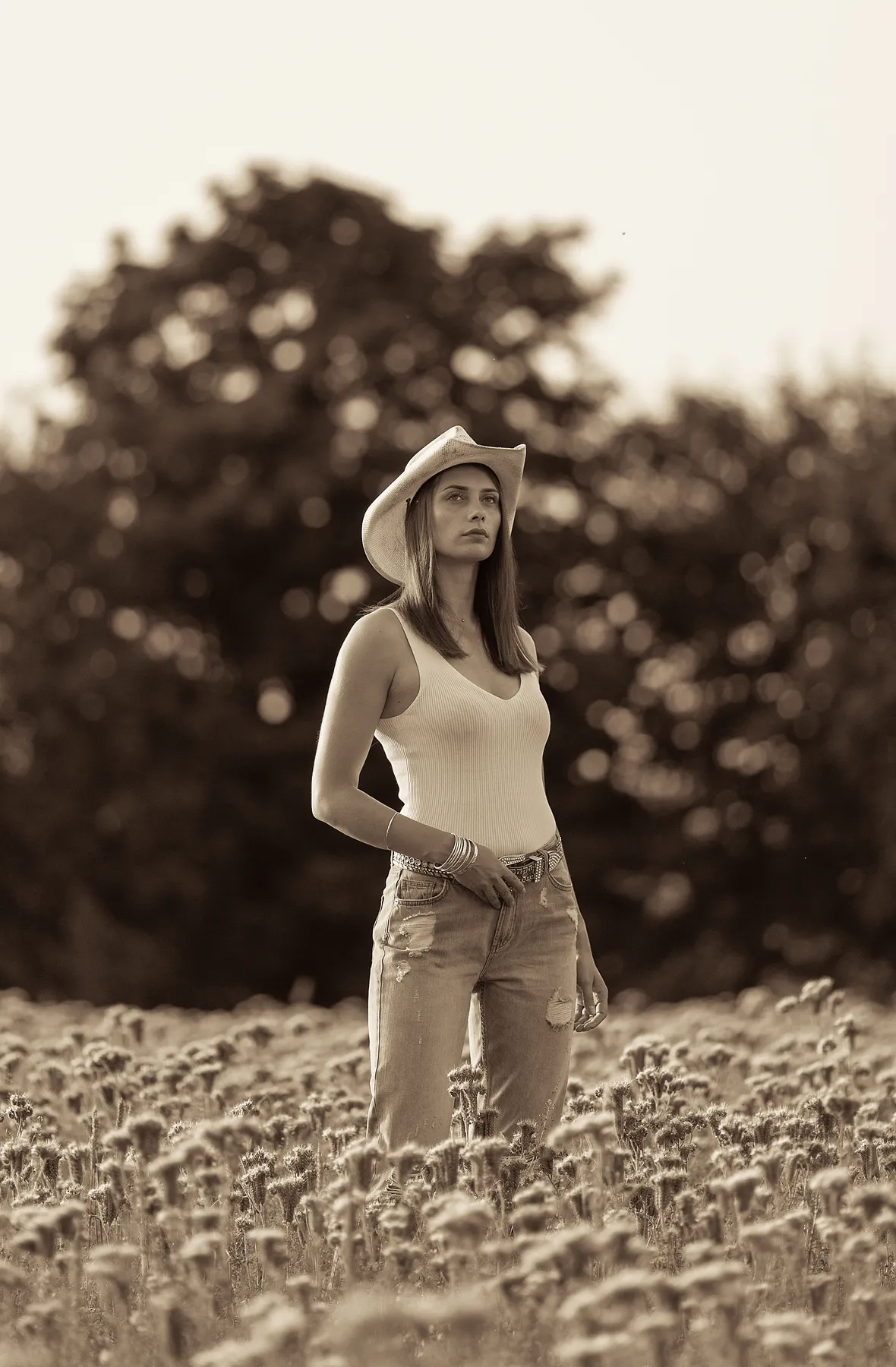 Eine Frau mit Cowboyhut, Tanktop und Jeans steht in einem Feld mit Wildblumen und Bäumen im Hintergrund. Das Bild ist in Sepiatönen gehalten.