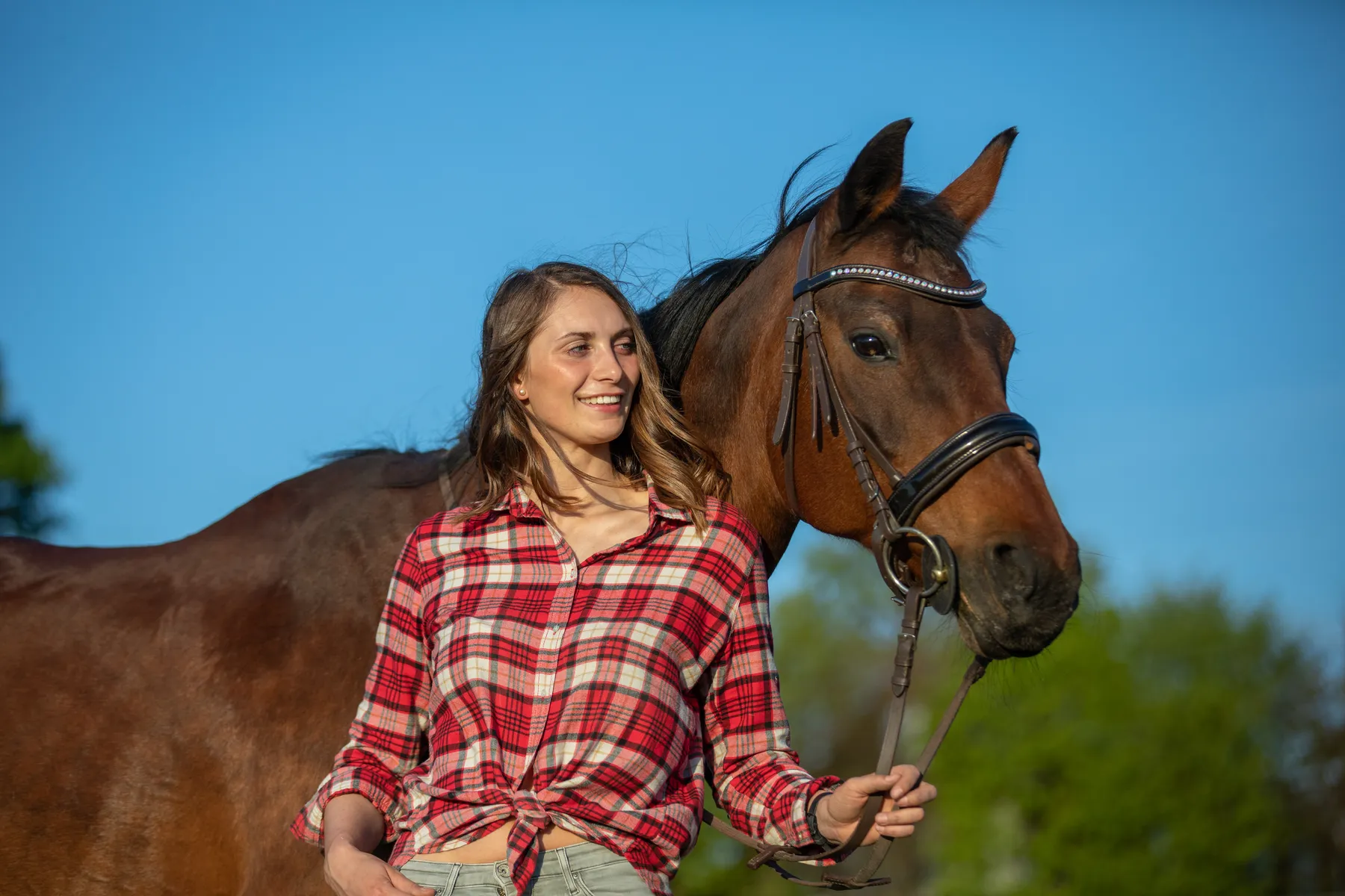 Eine Frau in einem rotkarierten Hemd hält die Zügel eines braunen Pferdes und lehnt mit ihrem Rücken dabei am Hals ihres Pferdes. Im Hintergrund sind grüne Bäume und ein blauer Himmel.