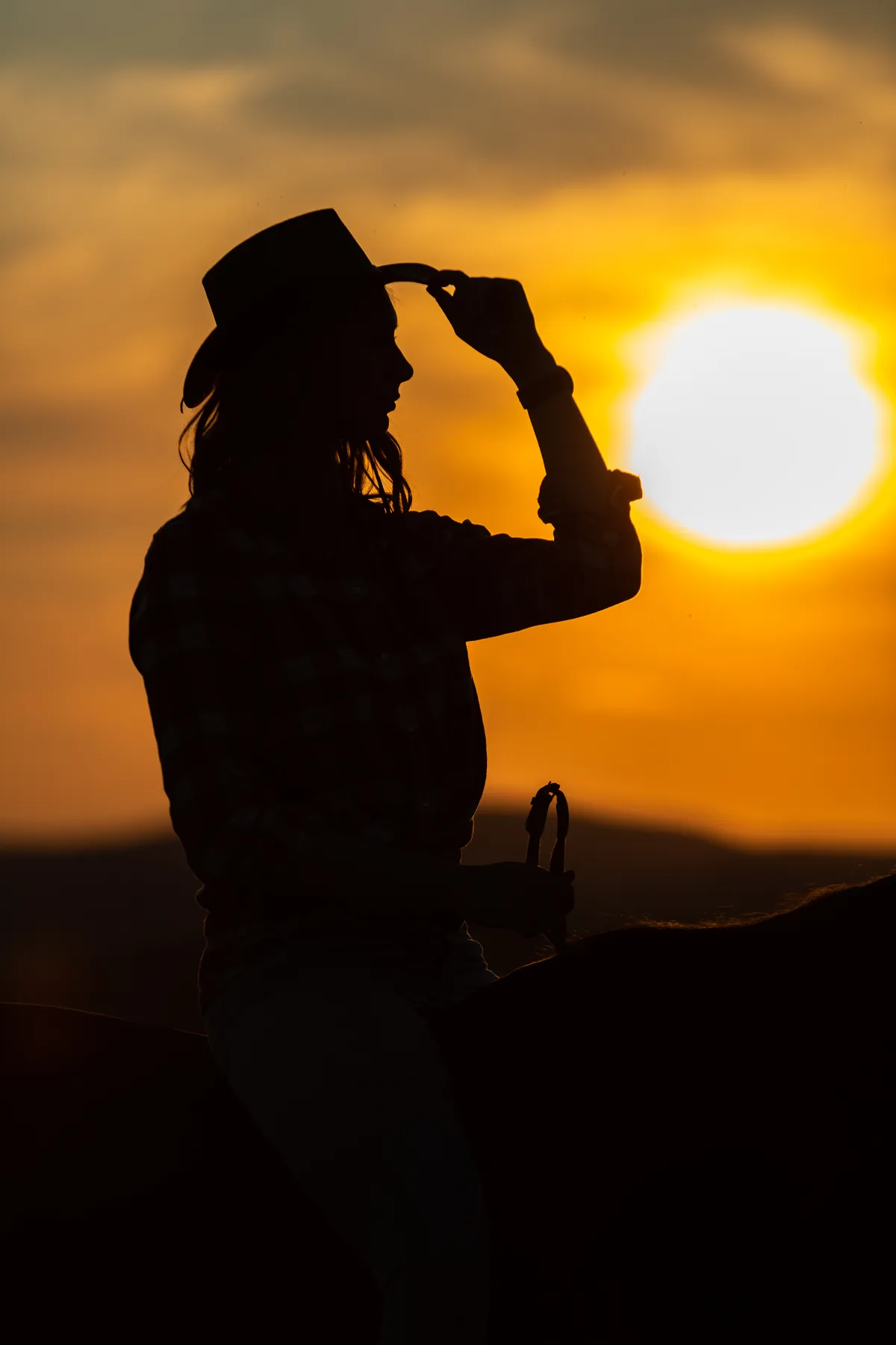 Silhouette einer Person mit Cowboyhut, die bei Sonnenuntergang auf einem Pferd sitzt, wobei die Sonne tief am Himmel steht und im Hintergrund orange-gelb leuchtet.