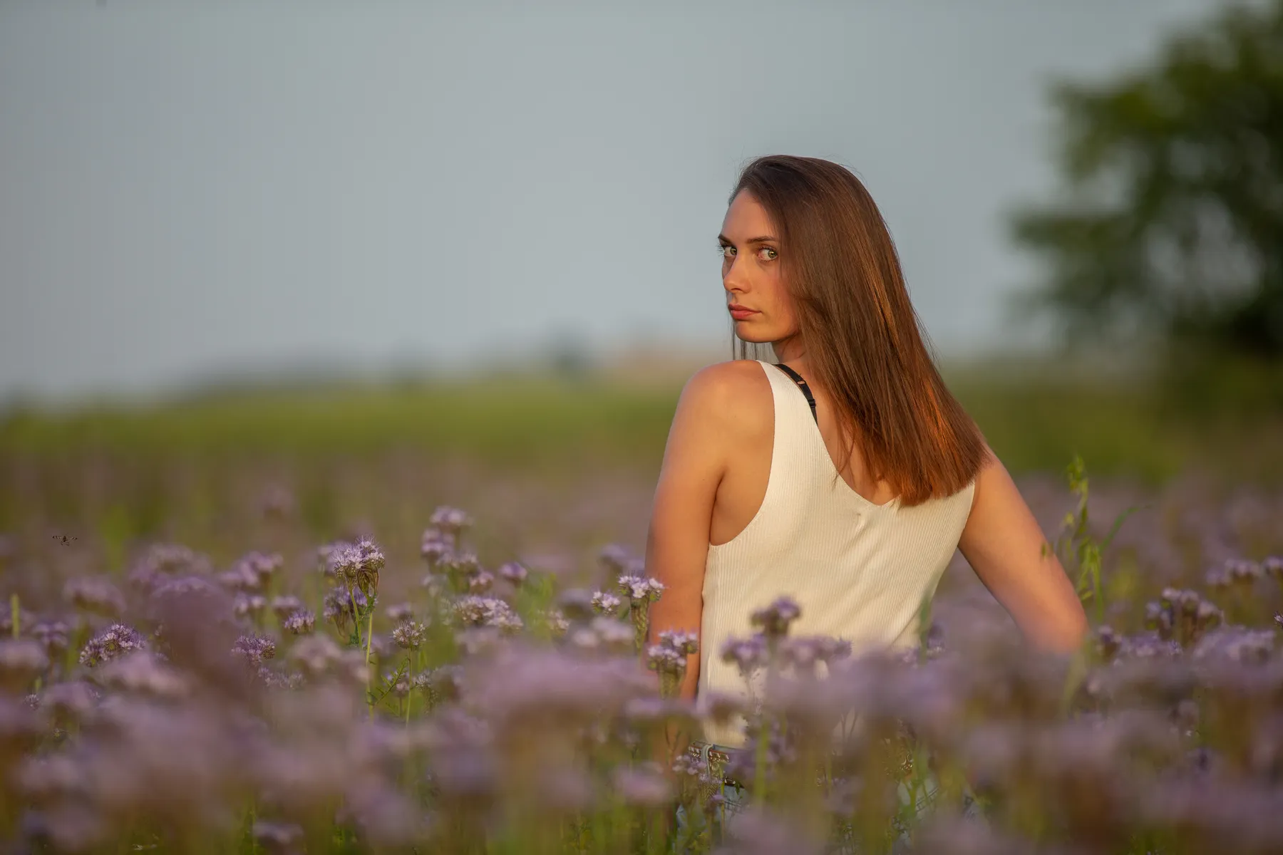 Eine Frau mit glattem braunem Haar und einem ärmellosen weißen Oberteil steht in einem Feld mit lila Blumen und blickt über ihre Schulter. Der Hintergrund ist unscharf, man sieht Grün und einen sanften Himmel.