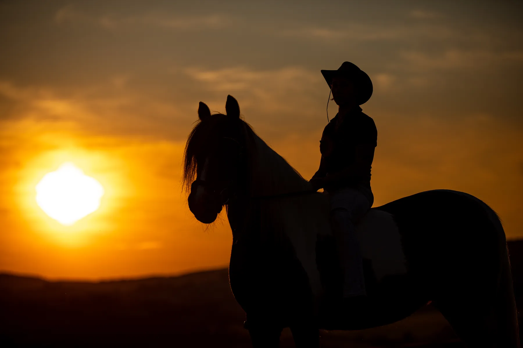 Eine Person mit Cowboyhut sitzt auf einem Pferd, beide sind vor einem orangefarbenen Sonnenuntergangshimmel zu sehen, wobei die Sonne tief am Horizont steht und die Berge im Hintergrund zu sehen sind.