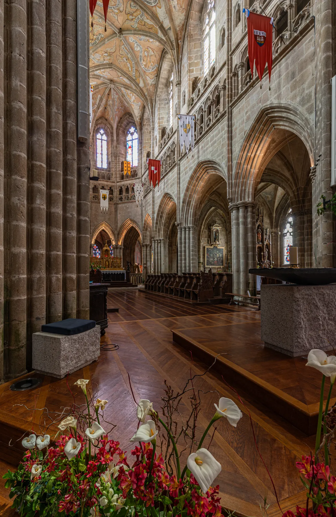 Verziertes gotisches Kathedraleninnere mit Gewölbedecken, Steinsäulen, roten Fahnen, hölzernem Chorgestühl und Blumenschmuck im Vordergrund. Das Sonnenlicht dringt durch die Buntglasfenster und erhellt die Szene.
