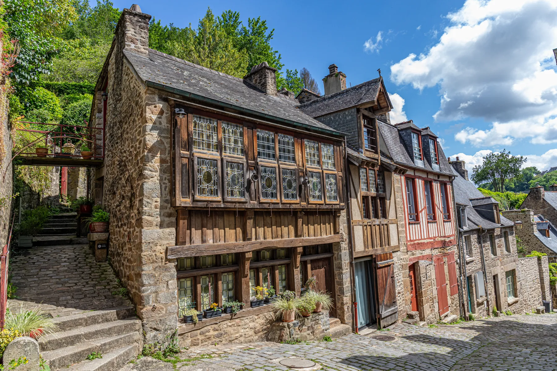 Landschaftsfotografie in der mittelalterlichen Stadt Dinan mit einer typischen Kopfsteinpflasterstraße und mit charmanten historischen Häusern aus Stein und Holz, von denen einige mit Blumenkästen geschmückt sind, und liegt mitten im Grünen unter einem blauen Himmel mit vereinzelten Wolken.