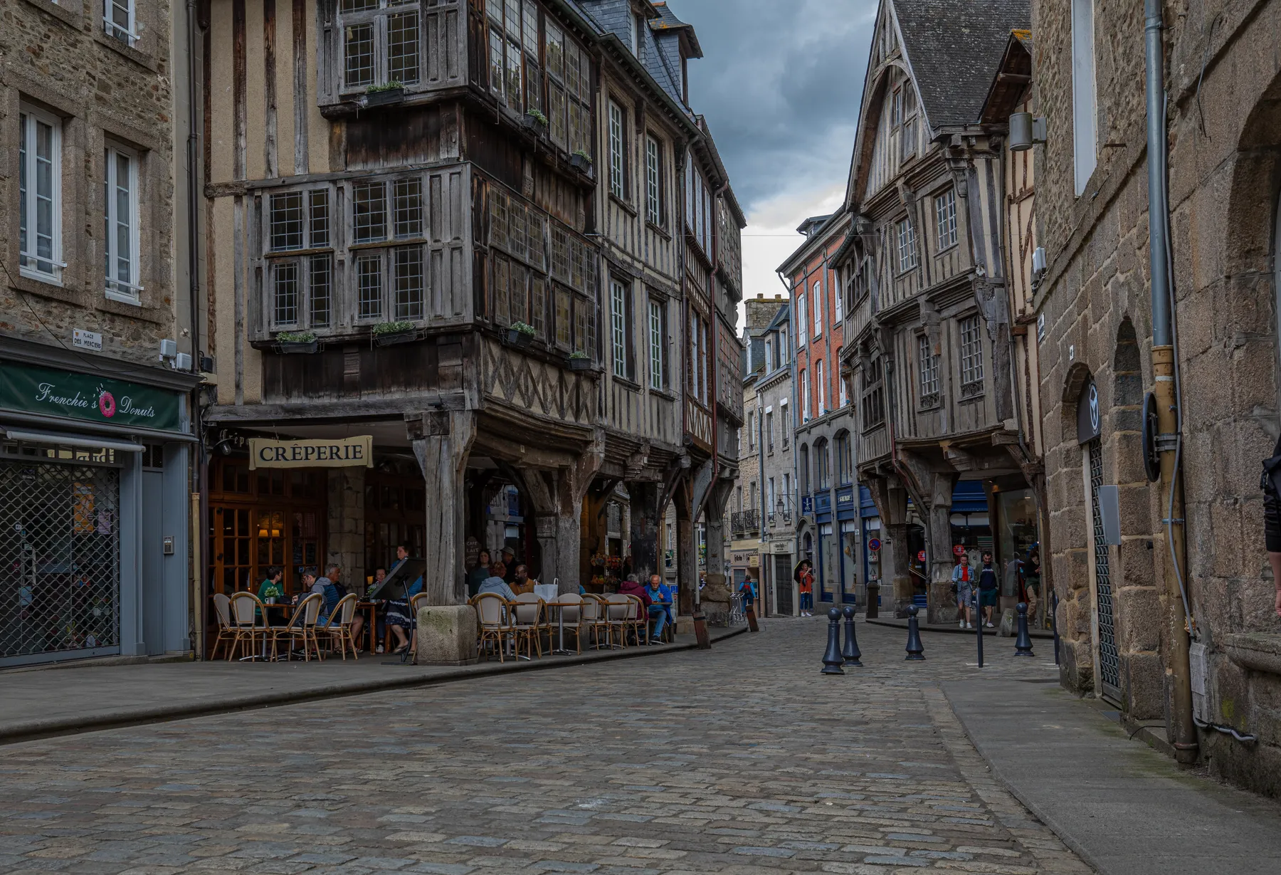 Eine kopfsteingepflasterte Straße in der Altstadt von Dinan, wo an einem bewölkten Tag Menschen in einer Crêperie im Freien unter Fachwerkhäusern speisen, umgeben von alter Steinarchitektur.