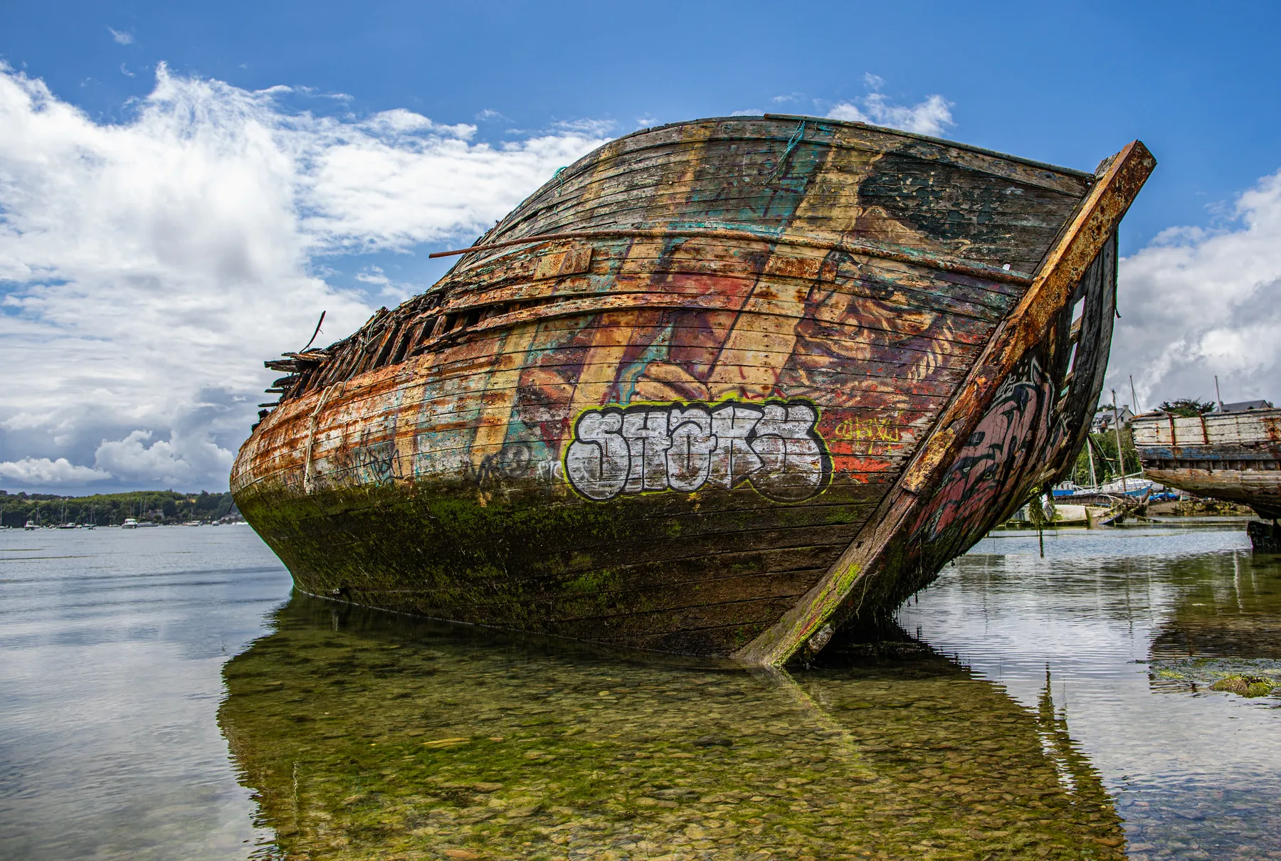 Landschaftsfotografie eines alten, verwitterten hölzernen Schiffswracks welches im flachen Wasser liegt, bedeckt mit bunten Graffiti, mit einem teilweise bewölkten Himmel und der Küstenlinie im Hintergrund.