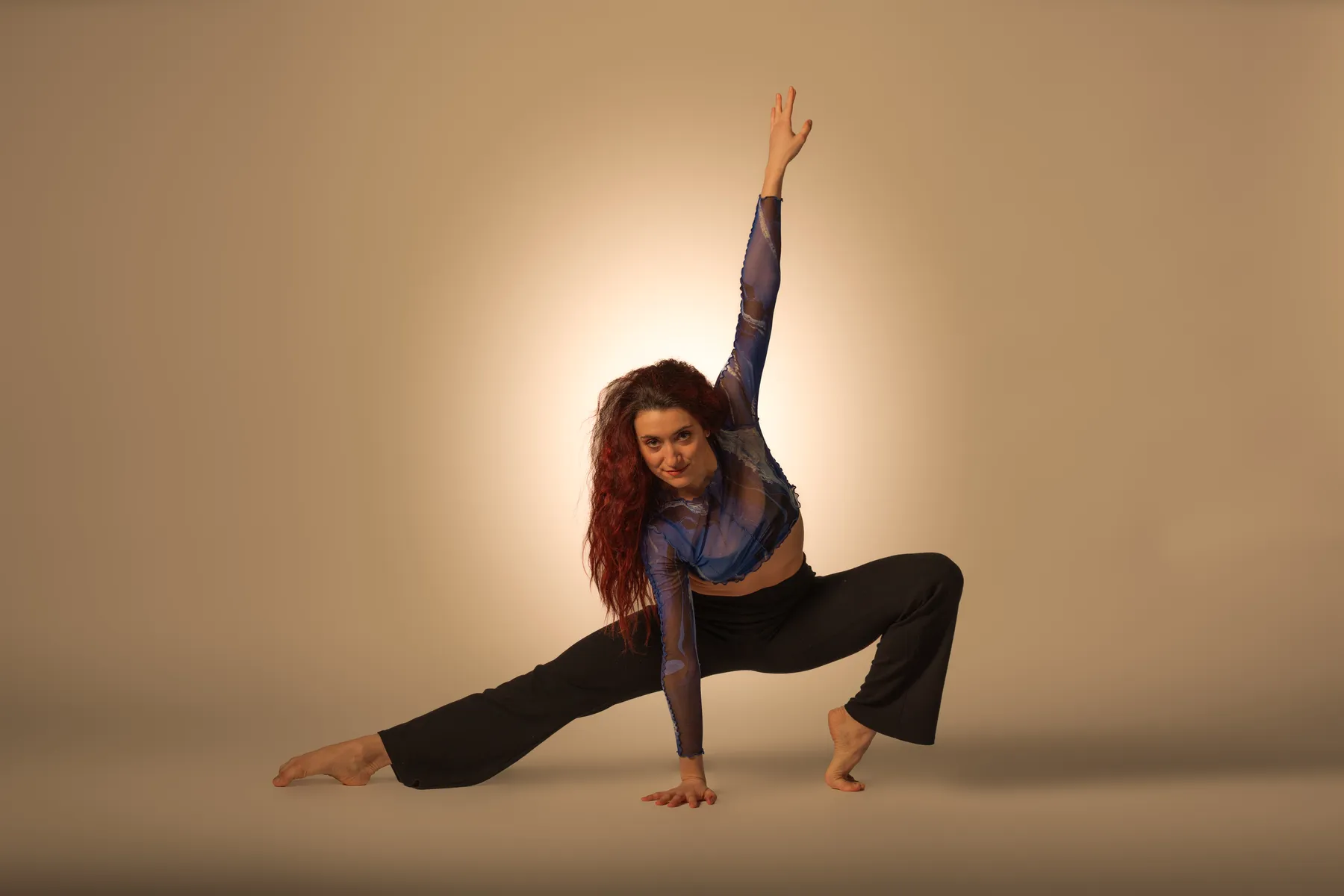 Zeitgenössische Tänzerin in blauer Transparenzbluse und schwarzer Hose in tiefer Ausfallschritt-Pose vor neutralem Hintergrund.