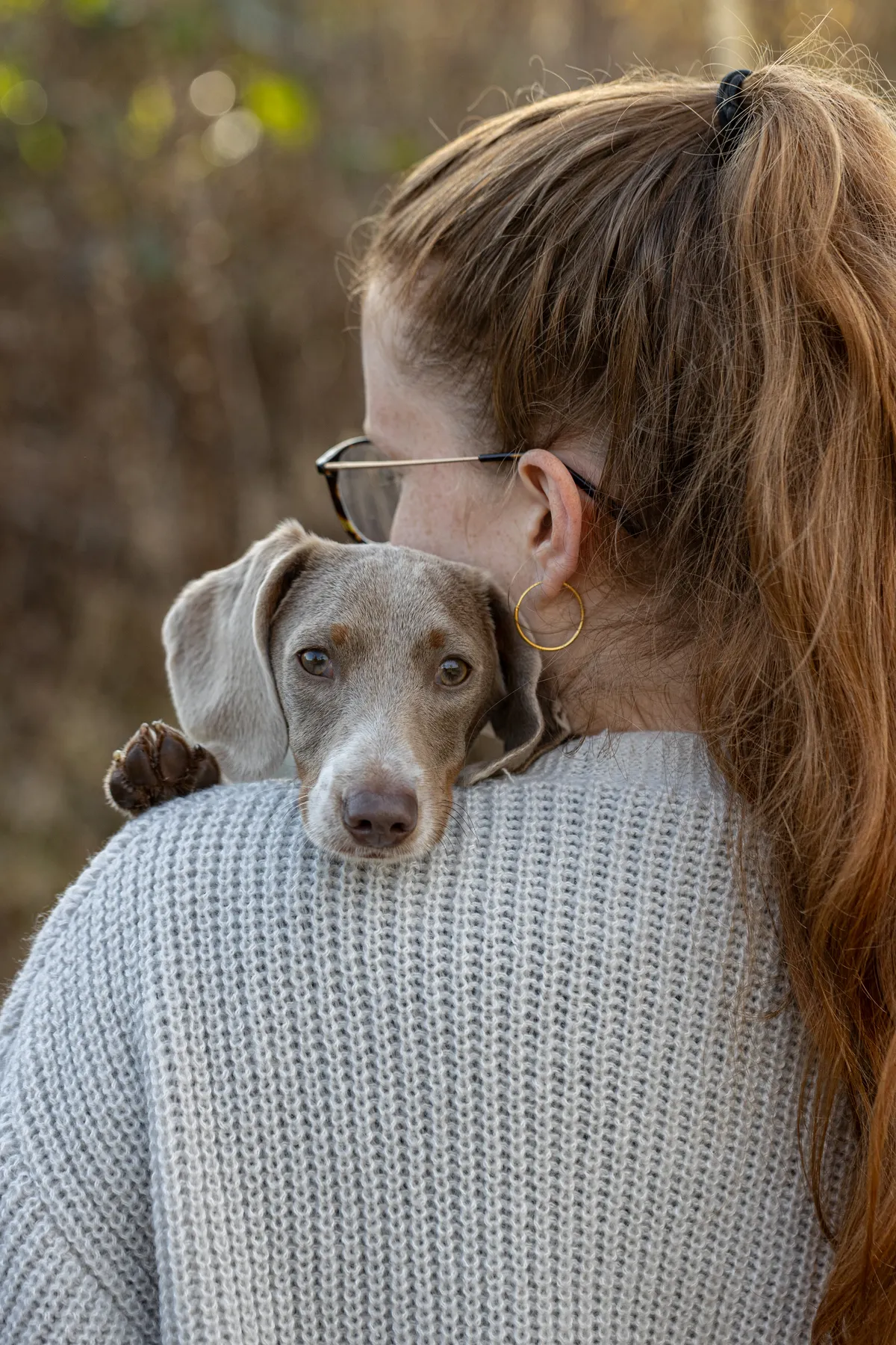 Eine Frau mit langen roten Haaren in einem Pferdeschwanz und einer Brille hält einen grauen Hund über ihre Schulter. Der Hund stützt sein Kinn und seine Pfote auf ihre Schulter und schaut in die Kamera. Sie trägt einen hellgrauen Strickpulli.