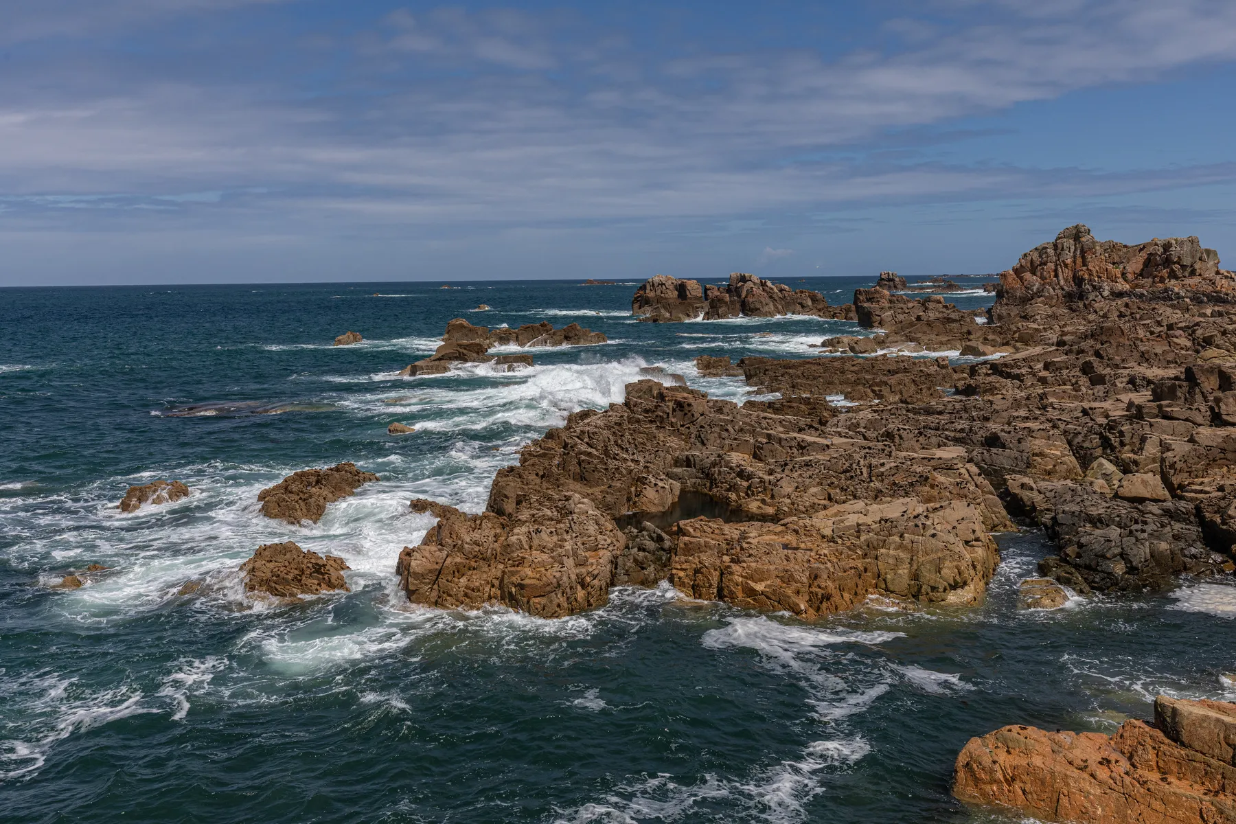 Die felsige Küste der Bretagne mit den Wellen, die gegen die zerklüfteten braunen Felsen schlagen, unter einem teilweise bewölkten Himmel und dem tiefblauen Wasser des Ozeans, das sich bis zum Horizont erstreckt.