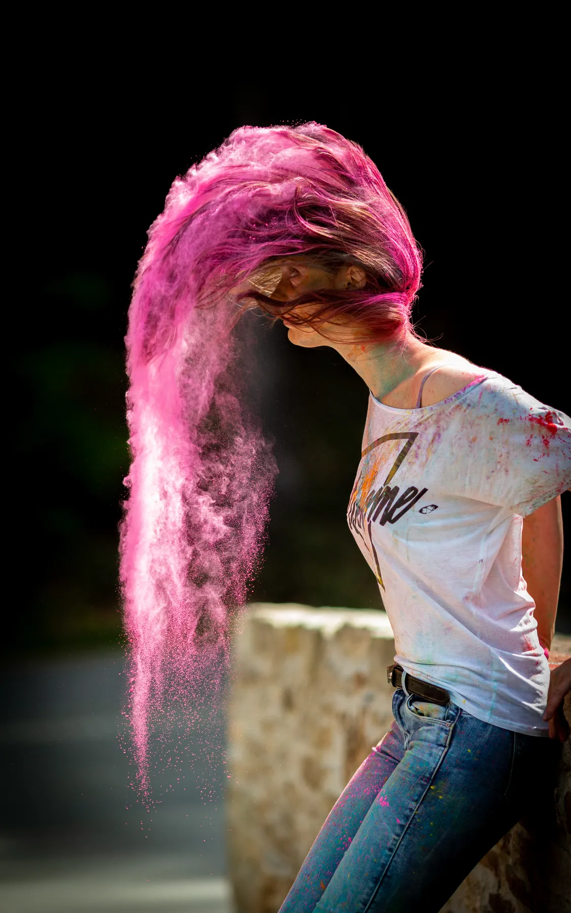 Eine Frau mit langen Haaren in weißem T-Shirt und Jeans hebt den Kopf und schickt eine Wolke aus leuchtendem Holi-Pulver in die Luft. Der Hintergrund ist dunkel, und die Frau lehnt an einer Steinmauer.