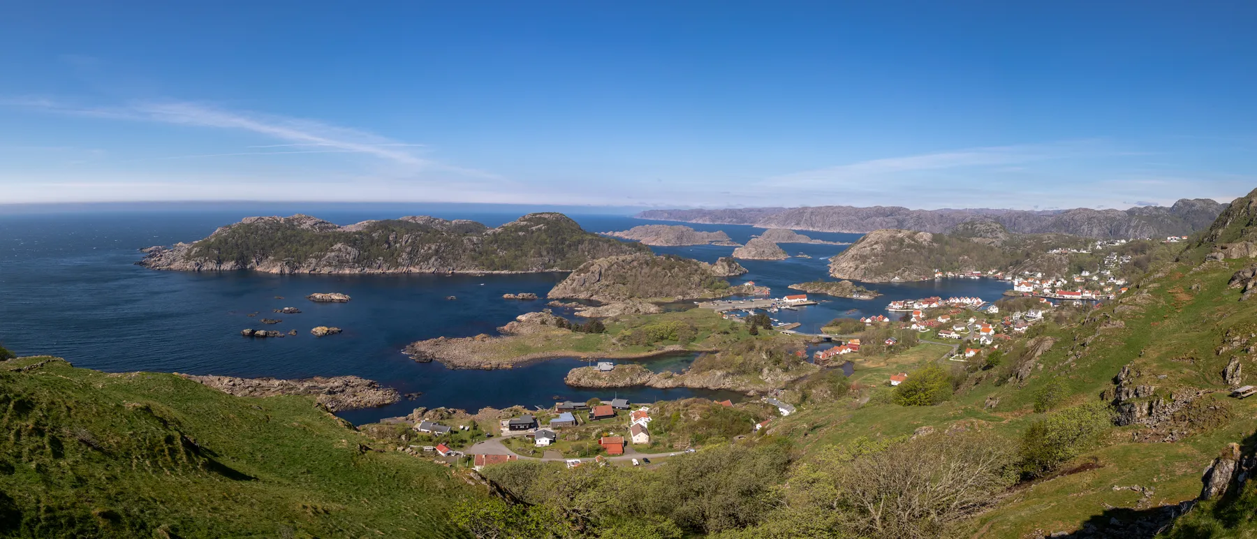 Landschaftsfotografie von Kirkehamn, ein Küstendorf mit Häusern, einem Jachthafen und grünen Hügeln, umgeben von felsigen Inseln und blauem Meer unter einem klaren Himmel.