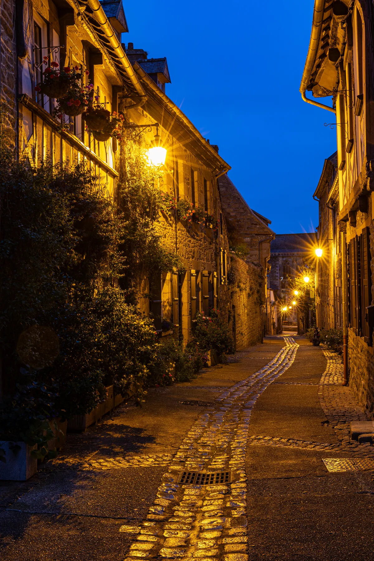 Eine schmale Kopfsteinpflasterstraße in Tréguier mit alten Steingebäuden wird in der Abenddämmerung von warmen Straßenlaternen beleuchtet, über denen ein tiefblauer Himmel hängt und auf der linken Seite etwas Grün zu sehen ist.