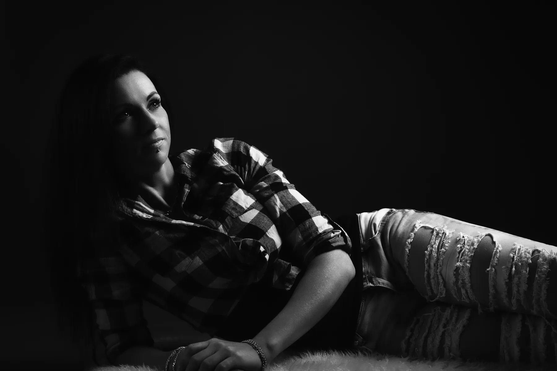 Eine Frau mit langen Haaren, einem karierten Hemd und zerrissenen Jeans lehnt sich in einer dunklen, unauffälligen Umgebung auf einer pelzigen Unterlage zurück und blickt mit neutralem Blick leicht nach oben. Die Aufnahme ist in dramatischer schwarz/ weiß Lichtsituation