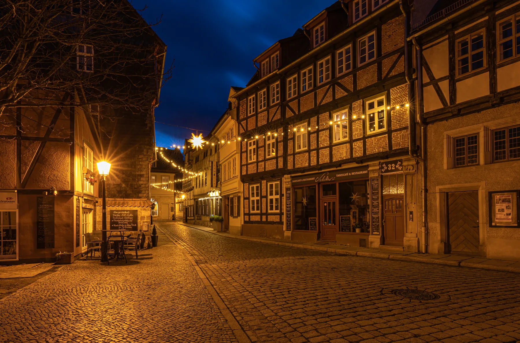 Kopfsteinpflasterstraße in Quedlinburg bei Nacht, gesäumt von Fachwerkhäusern, warmen Straßenlaternen und festlichen Lichterketten, die eine gemütliche, einladende Atmosphäre schaffen. Es sind keine Menschen zu sehen.