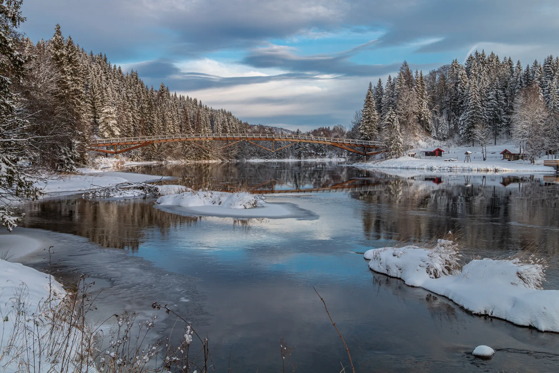 Eine verschneite Winterlandschaft im Kjærra Foss Park, mit einem Fluss, in dem sich Bäume und ein bewölkter Himmel spiegeln, einer Holzbrücke über den Fluß Lågen und roten Hütten zwischen schneebedeckten Tannen auf der rechten Seite.