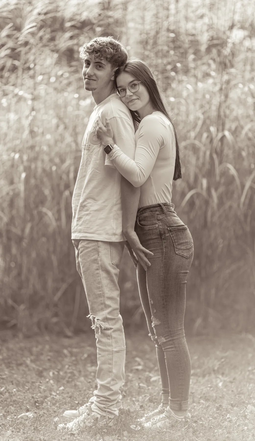 Ein junger Mann und eine Frau stehen im Freien dicht beieinander. Die Frau umarmt den Mann von hinten und beide schauen in die Kamera. Sie sind leger gekleidet und stehen auf einer Wiese mit hohen Pflanzen im Hintergrund. Das Bild ist in Sepiatönen gehalten.
