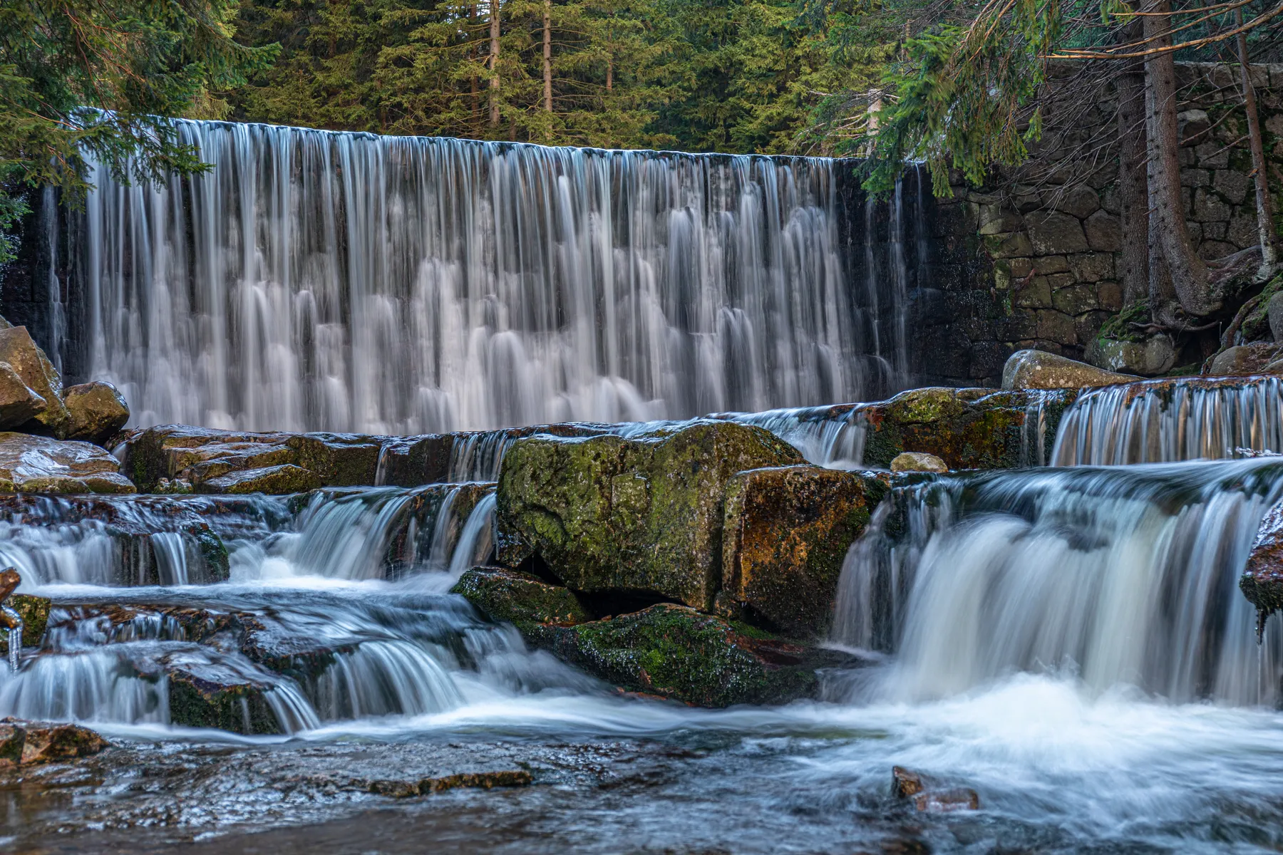 Ein ruhiger Wasserfall stürzt über einen Steindamm in einen felsigen Bach, umgeben von grünen Bäumen und moosbewachsenen Felsbrocken in einer Waldlandschaft.