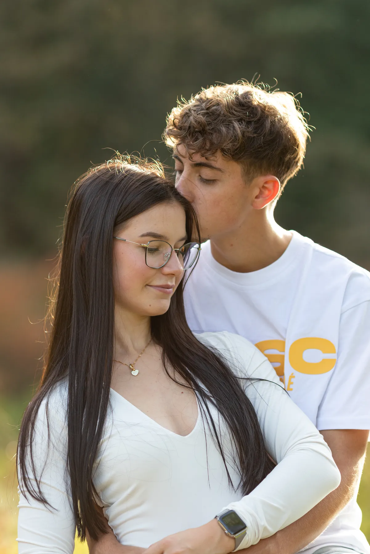 Ein junger Mann in einem weißen T-Shirt küsst sanft die Stirn einer jungen Frau mit langen dunklen Haaren und Brille, die ein weißes Top und eine Smartwatch trägt. Sie befinden sich im Freien vor einem unscharfen natürlichen Hintergrund.