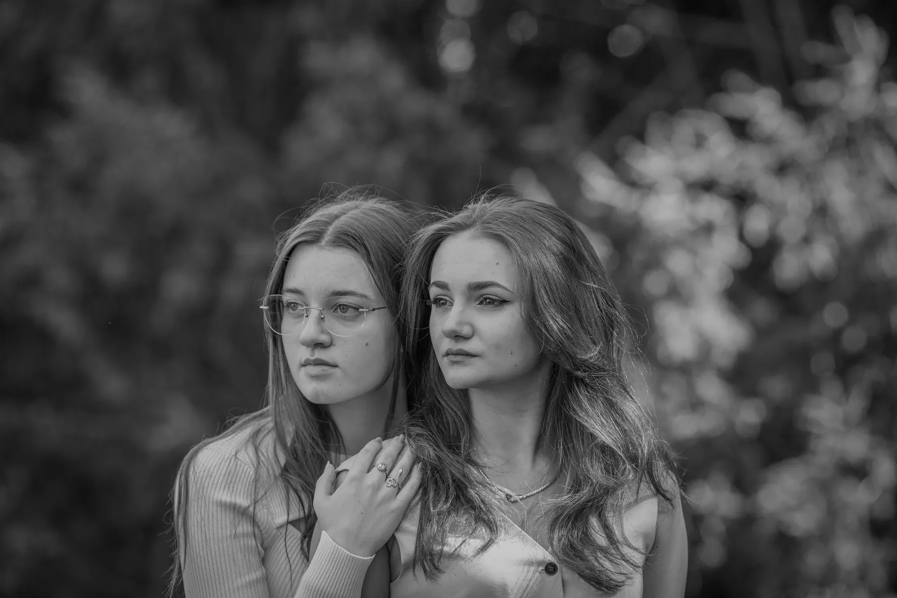 Zwei junge Frauen stehen im Freien dicht beieinander, eine mit Brille und glattem Haar, die andere mit gewelltem Haar. Die junge Frau mit Brille steht hinter der anderen und hat ihre Hand auf ihre Schulter gelegt. Sie blicken mit ernster Miene in die gleiche Richtung. Der Hintergrund besteht aus unscharfem Laub.