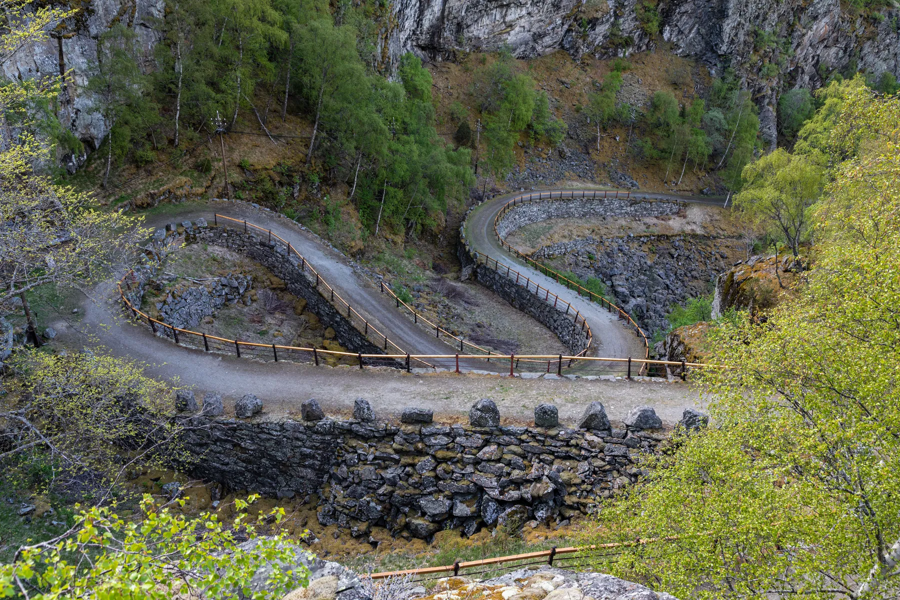 Ein kurvenreicher Bergpfad, der Vindhellavegen, mit scharfen Serpentinen, gesäumt von Holzgeländern, schlängelt sich durch felsiges Gelände und grüne Bäume, mit steilen Klippen und bewaldeten Gebieten im Hintergrund.