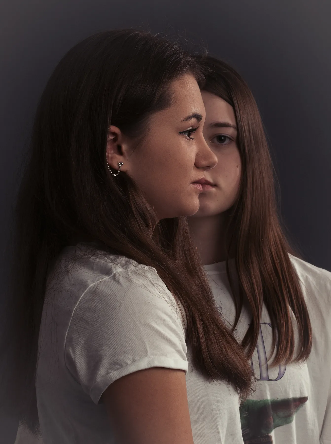 Zwei junge Frauen mit langen braunen Haaren stehen dicht nebeneinander vor einem dunklen Hintergrund. Die eine steht im Profil nach links, während die andere hinter ihr steht und nach vorne schaut. Es sind zwei halbe Gesichter zu sehen die durch die Stellung wieder eines ergeben, aber aus zwei Personen. Beide tragen weiße T-Shirts. Die Aufnahme ist im Studio.