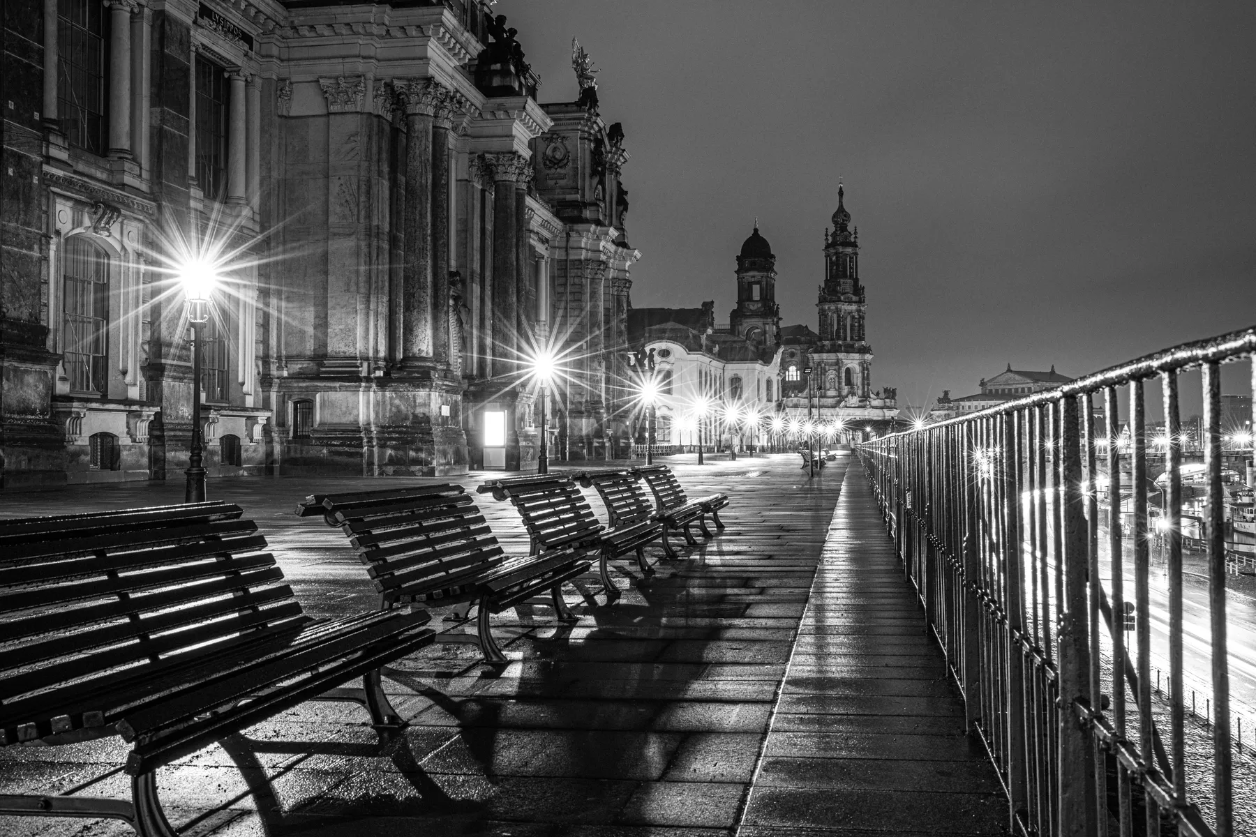 Ein Schwarz-Weiß-Foto von leeren Bänken auf der nassen, beleuchteten Brühlschen Terrasse bei Dresden, mit historischen Gebäuden und Kirchtürmen im Hintergrund, während sich helle Straßenlaternen auf dem Pflaster spiegeln.