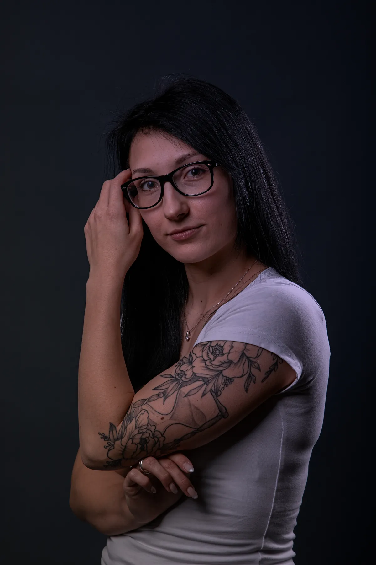 Eine Frau mit langen dunklen Haaren und Brille steht vor einem dunklen Hintergrund und trägt ein weißes T-Shirt. Sie hat auf ihrem linken Oberarm eine Blumentätowierung und schaut in die Kamera, während sie mit der rechten Hand ihre Brille berührt.