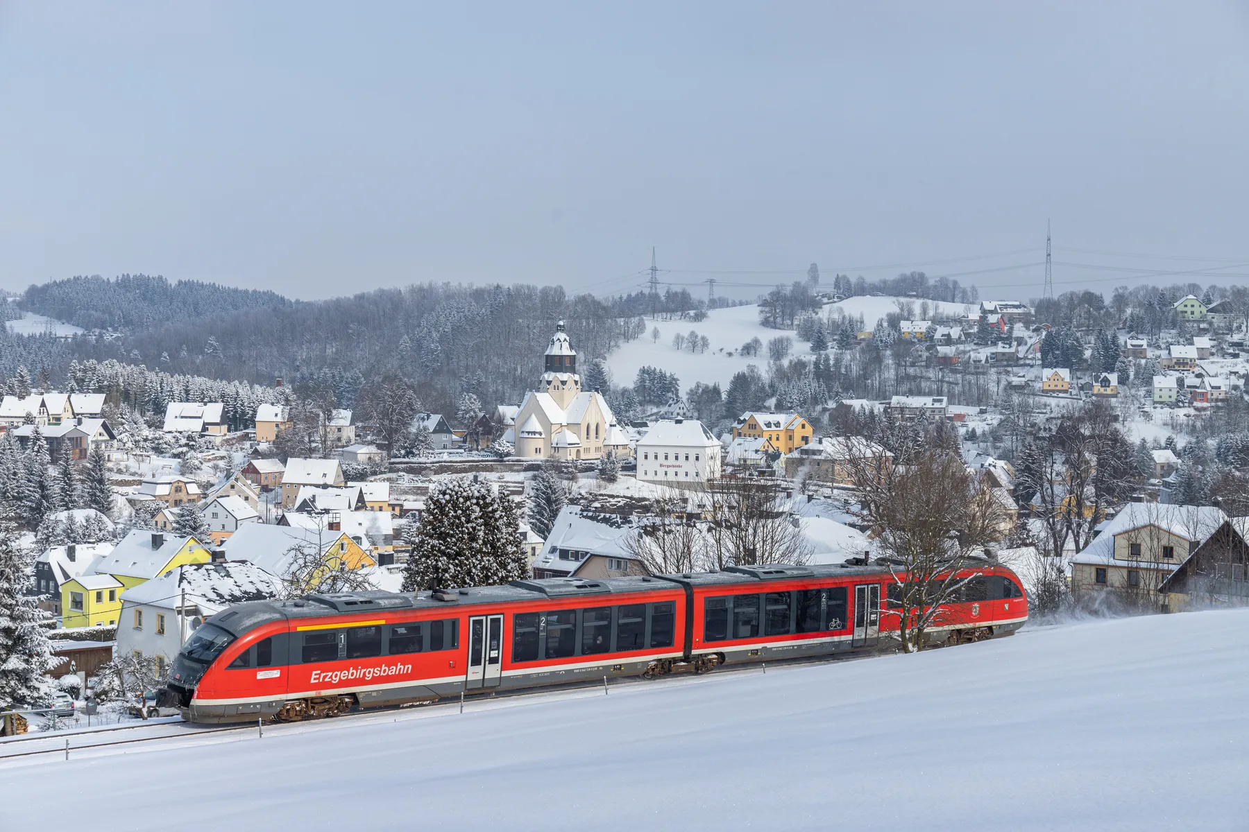 Erzgebirgsbahn fährt durch das verschneite Wiesa mit der St.-Trinitatis-Kirche im Hintergrund