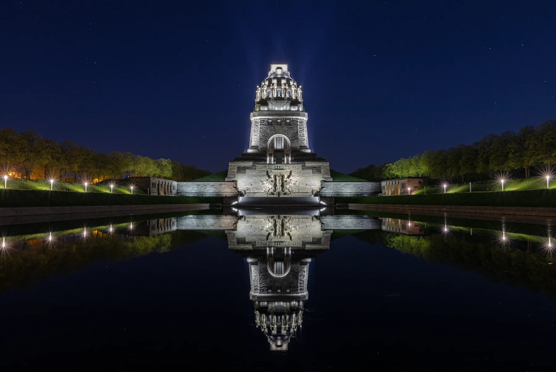 Landschaftsfotografie vom Völkerschlachtdenkmal, ein großes beleuchtetes Steinmonument in Leipzig, spiegelt sich nachts im ruhigen Wasser, umgeben von Bäumen und beleuchteten Wegen unter einem klaren Sternenhimmel.