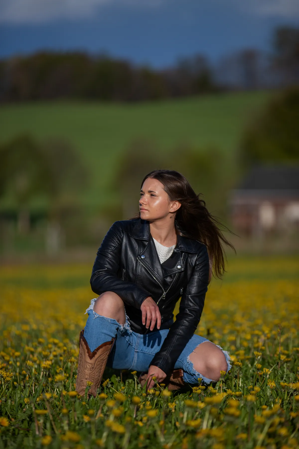 Eine Frau in schwarzer Lederjacke, Jeansshorts und Cowboystiefeln kniet in einem Feld mit gelben Blumen vor einem unscharfen Hintergrund mit grünen Hügeln, Bäumen und einem Gebäude. Der Wind weht sanft ihr Haar nach hinten