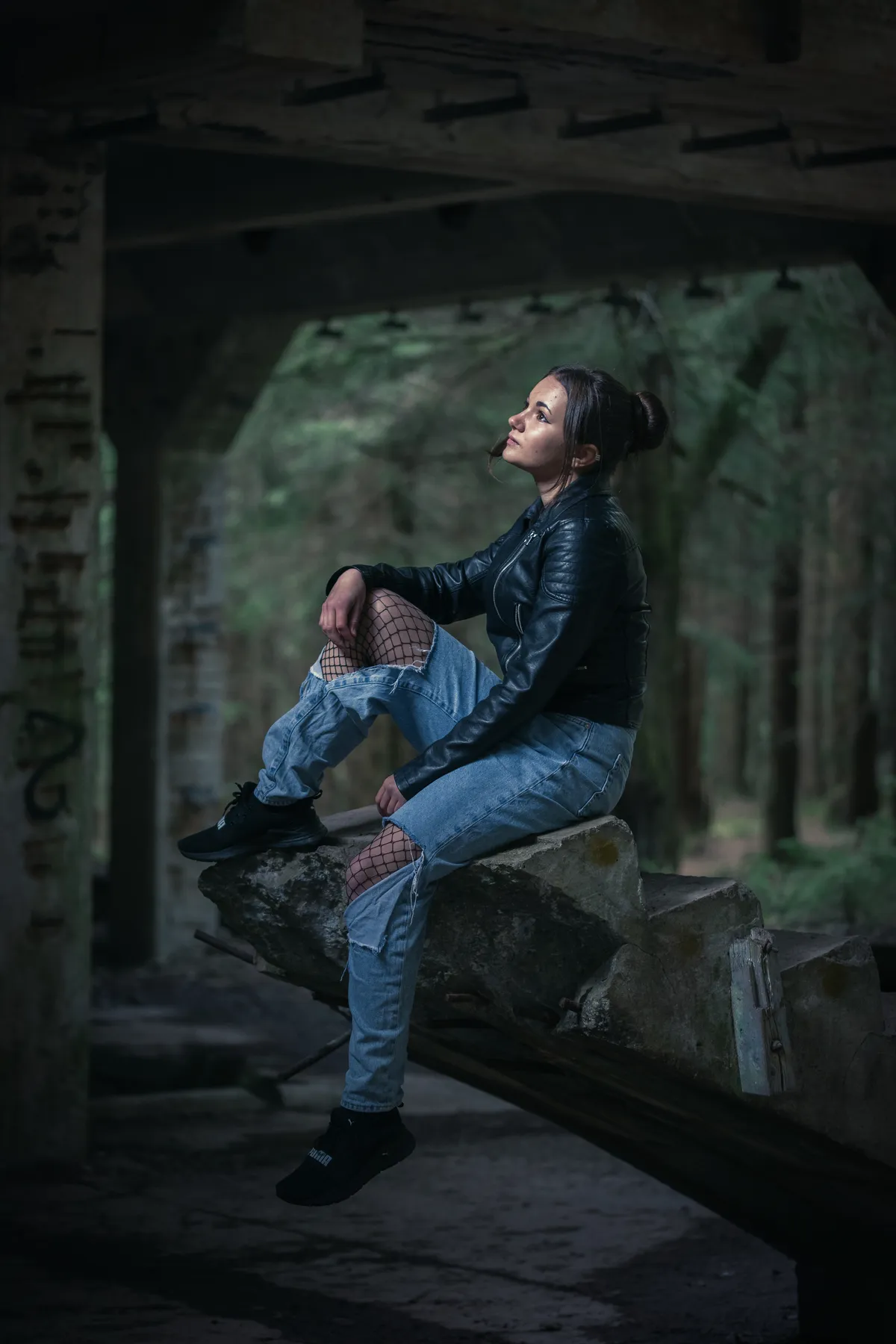 Eine Person, die eine schwarze Lederjacke, zerrissene Jeans mit Netzstrumpfhose und schwarze Schuhe trägt, sitzt auf einer großen abgebrochenen Treppe in einem verlassenen Gebäude, das von Bäumen umgeben ist, und blickt nach oben.
