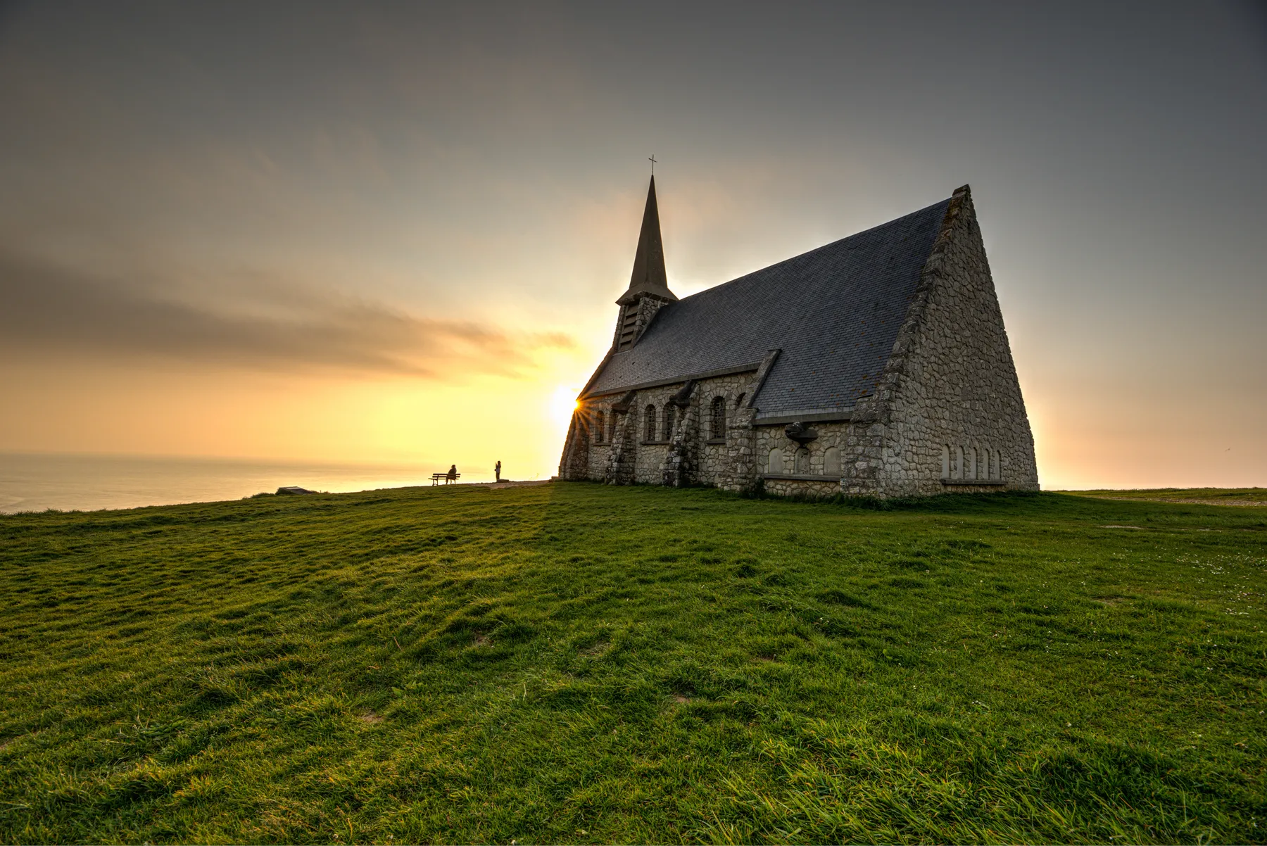 Eine Steinkapelle in Étretat mit einem Kirchturm steht auf einem grasbewachsenen Hügel bei Sonnenuntergang. Die Sonne geht hinter dem Gebäude unter und wirft ein warmes Licht. Zwei Menschen und eine Bank zeichnen sich in der Nähe des Horizonts ab.