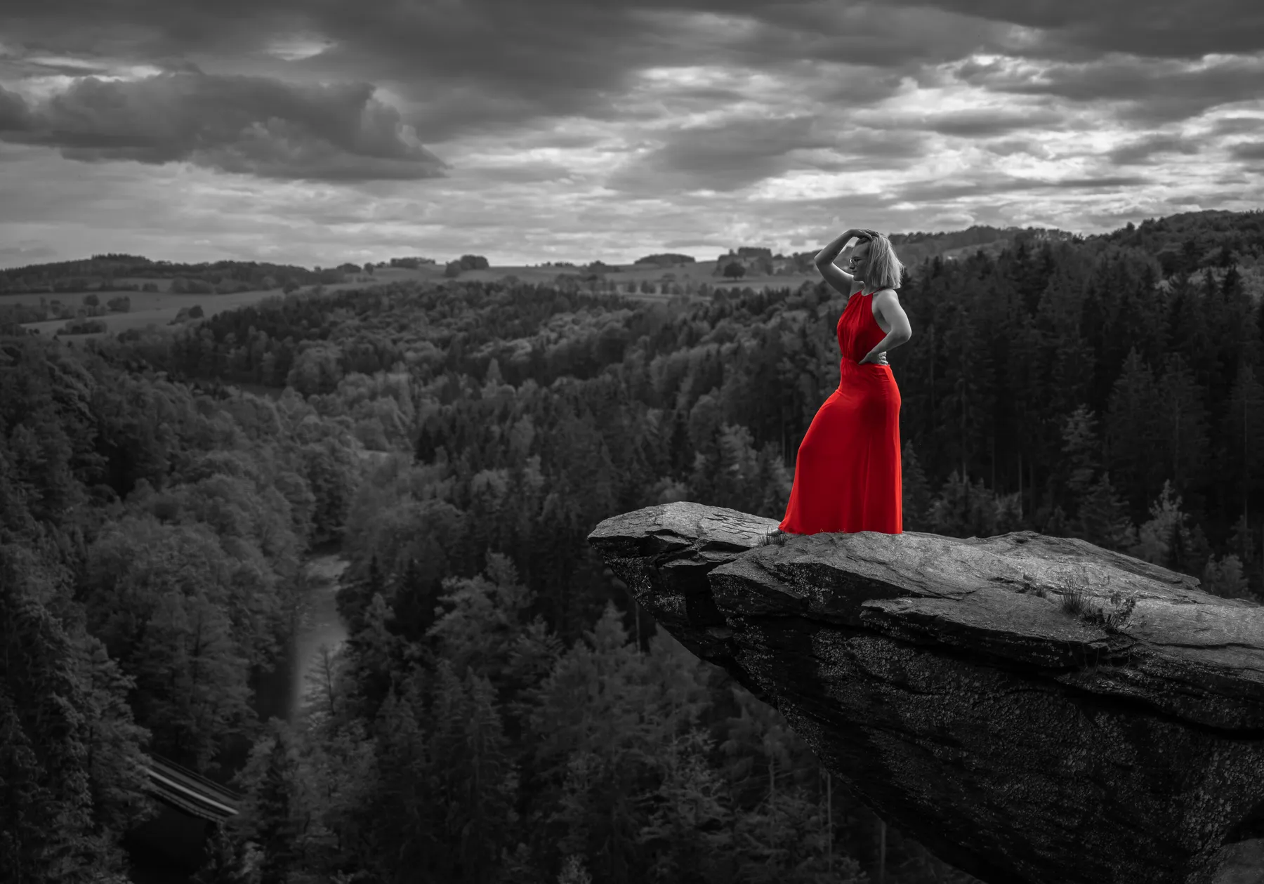Eine Frau in einem leuchtend roten Kleid steht am Rande einer felsigen Klippe mit Blick auf einen großen Wald und einen Fluss unter einem bewölkten Himmel. Der Hintergrund ist in Schwarz und Weiß gehalten, wodurch das rote Kleid hervorgehoben wird.