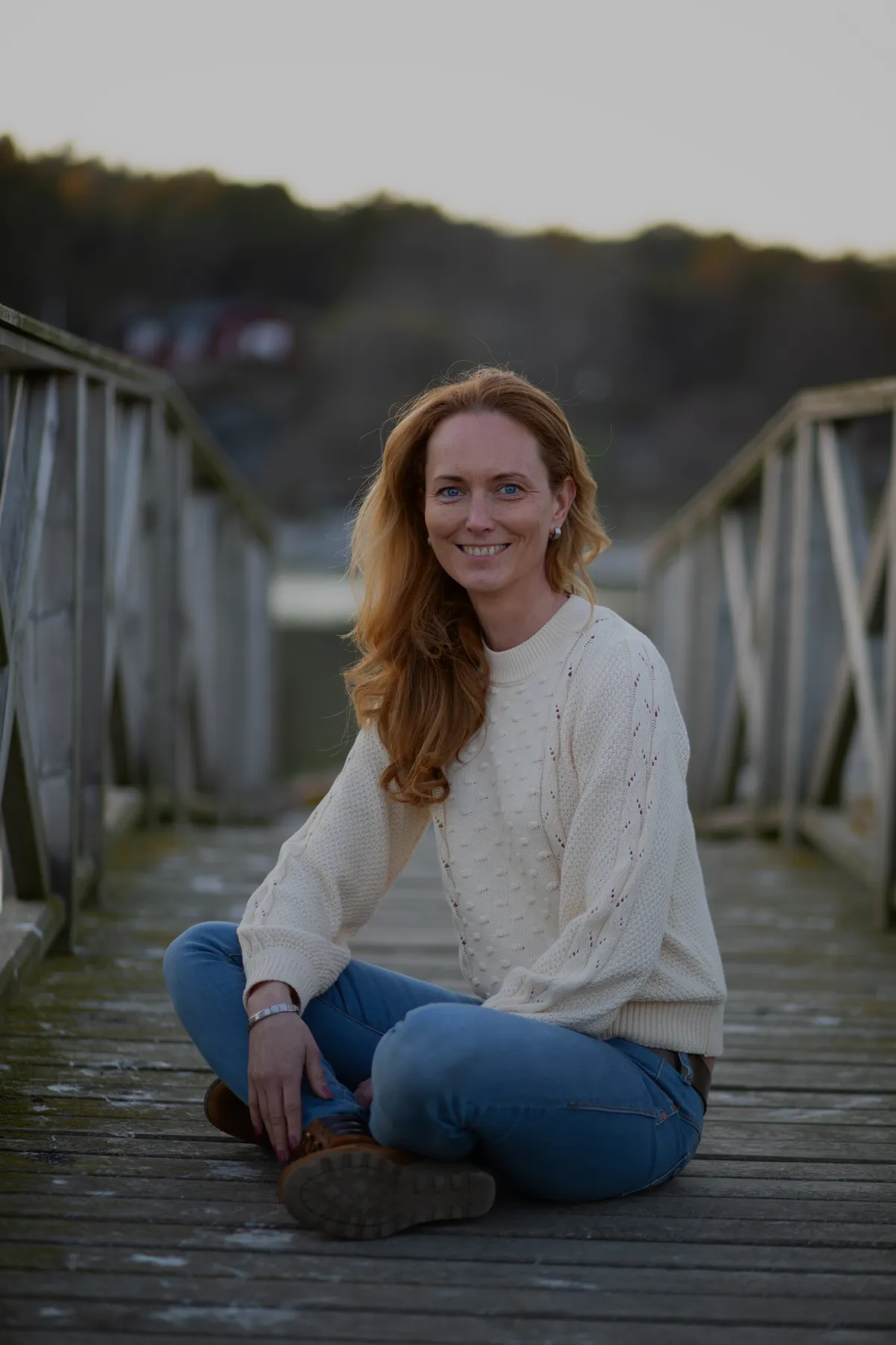 Eine Frau mit langen dunkelblonden Haaren, die einen cremefarbenen Pullover und blaue Jeans trägt, sitzt im Schneidersitz auf einer Holzbrücke im Freien und lächelt in die Kamera. Im Hintergrund sind Bäume und verschwommene Häuser zu sehen.