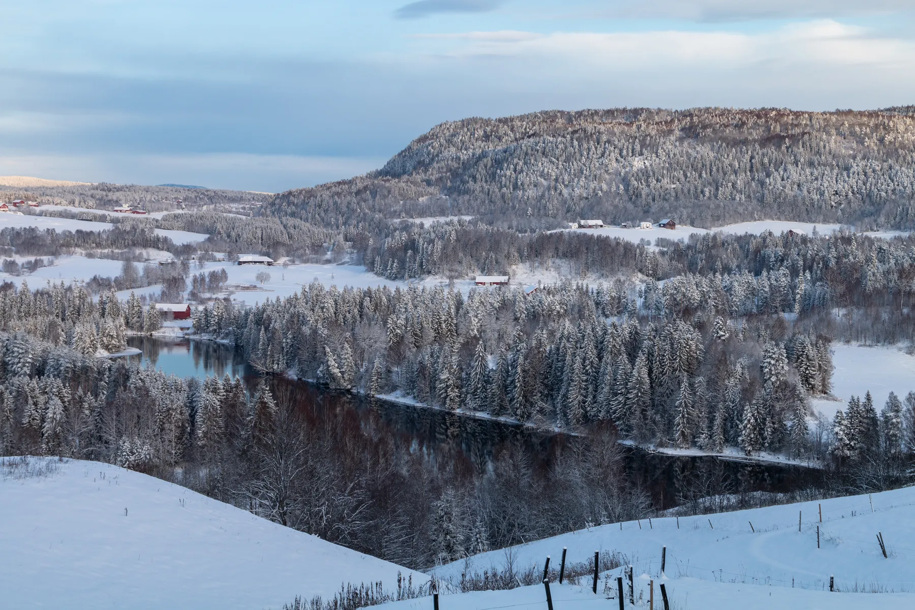 Landschaftsfotografie einer verschneiten norwegischen Landschaft mit einem sich durch Nadelwäldern schlängelnden Fluss und verstreuten Häusern, umgeben von schneebedeckten Hügeln und Bäumen unter einem teilweise bewölkten Himmel.