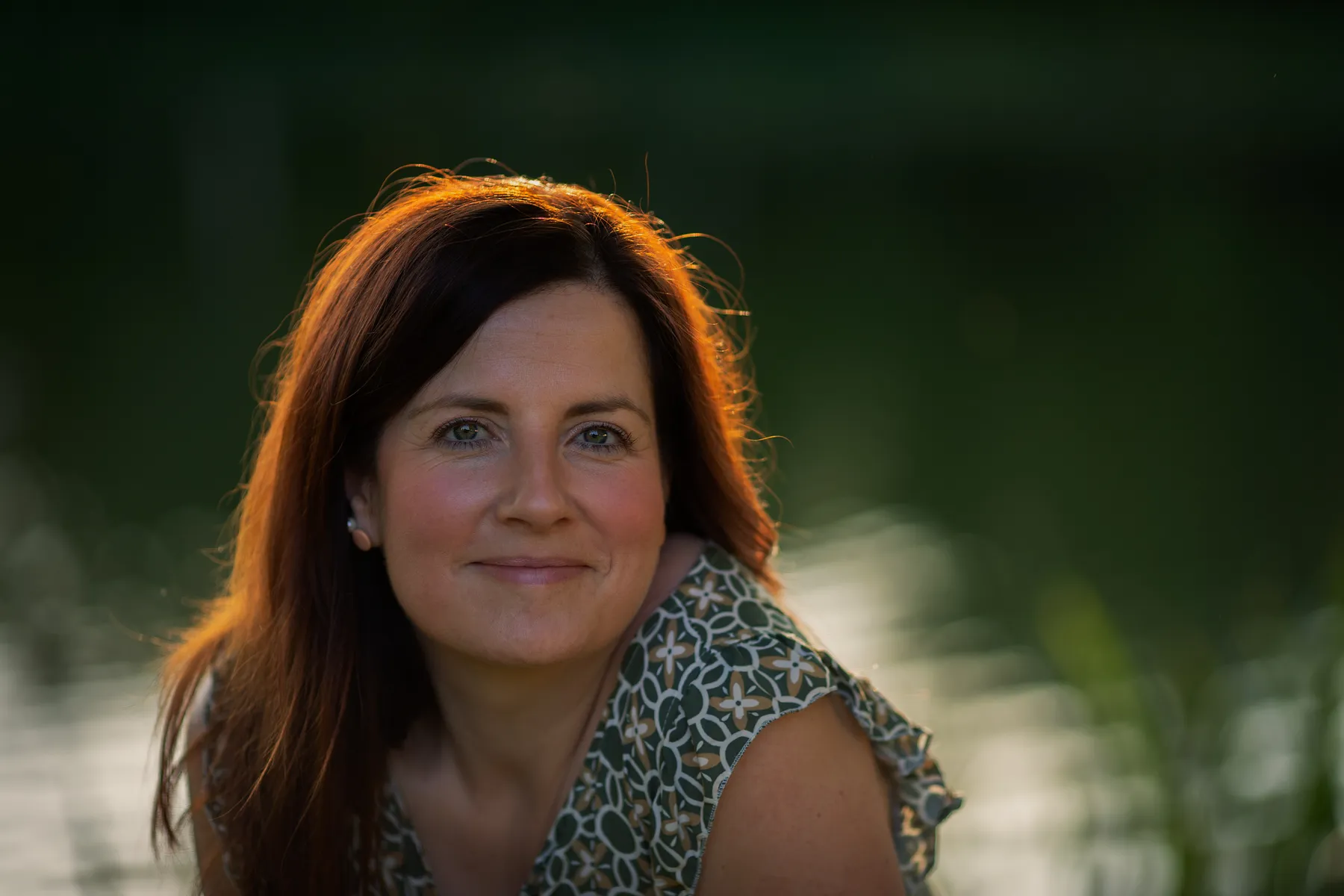 Portraitfotografie einer lächelnden Frau mit braunen Haaren in natürlicher Umgebung am Wasser, aufgenommen bei warmem Abendlicht mit weichem, unscharfem Hintergrund.