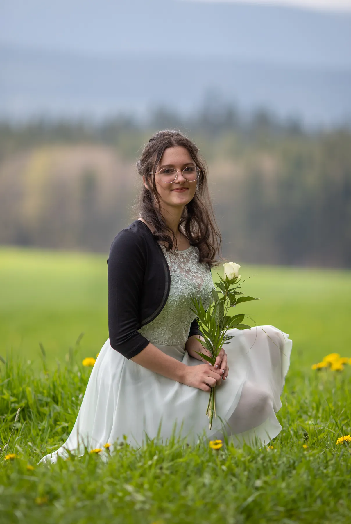 Junges Mädchen kniet auf grüner Wiese, hält weiße Rose, trägt festliches Kleid und Brille, lächelt sanft, natürliche Landschaft im Hintergrund, Konfirmation Portraitfotograf Aufnahme bei weichem Licht draußen Frühling ruhig elegant