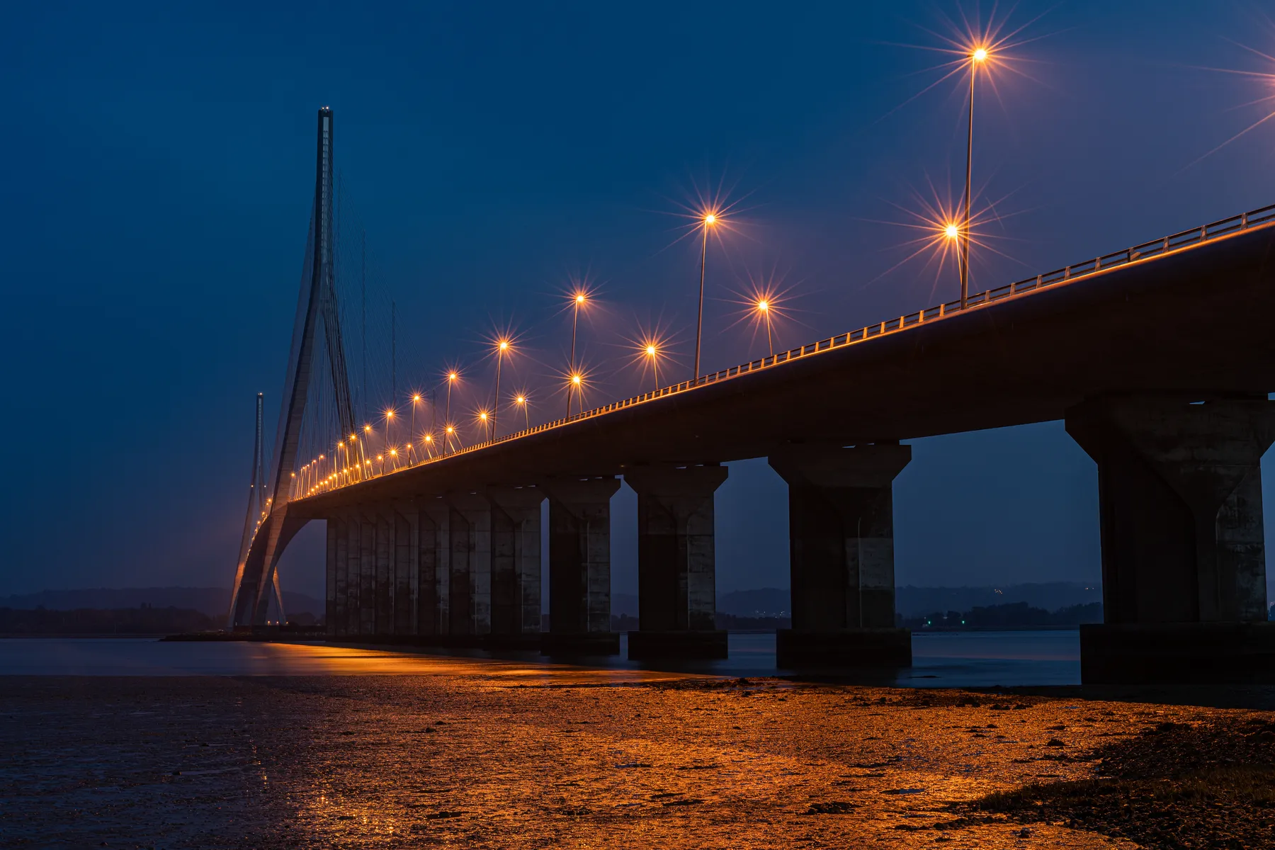 Die große, beleuchtete Schrägseilbrücke Pont de Normandie spannt sich nachts über das ruhige Wasser. Die orangefarbenen Straßenlaternen spiegeln sich auf dem Wasser und dem nassen Ufer unter ihr. Der Himmel ist tiefblau, und die Türme der Brücke ragen in die Ferne.