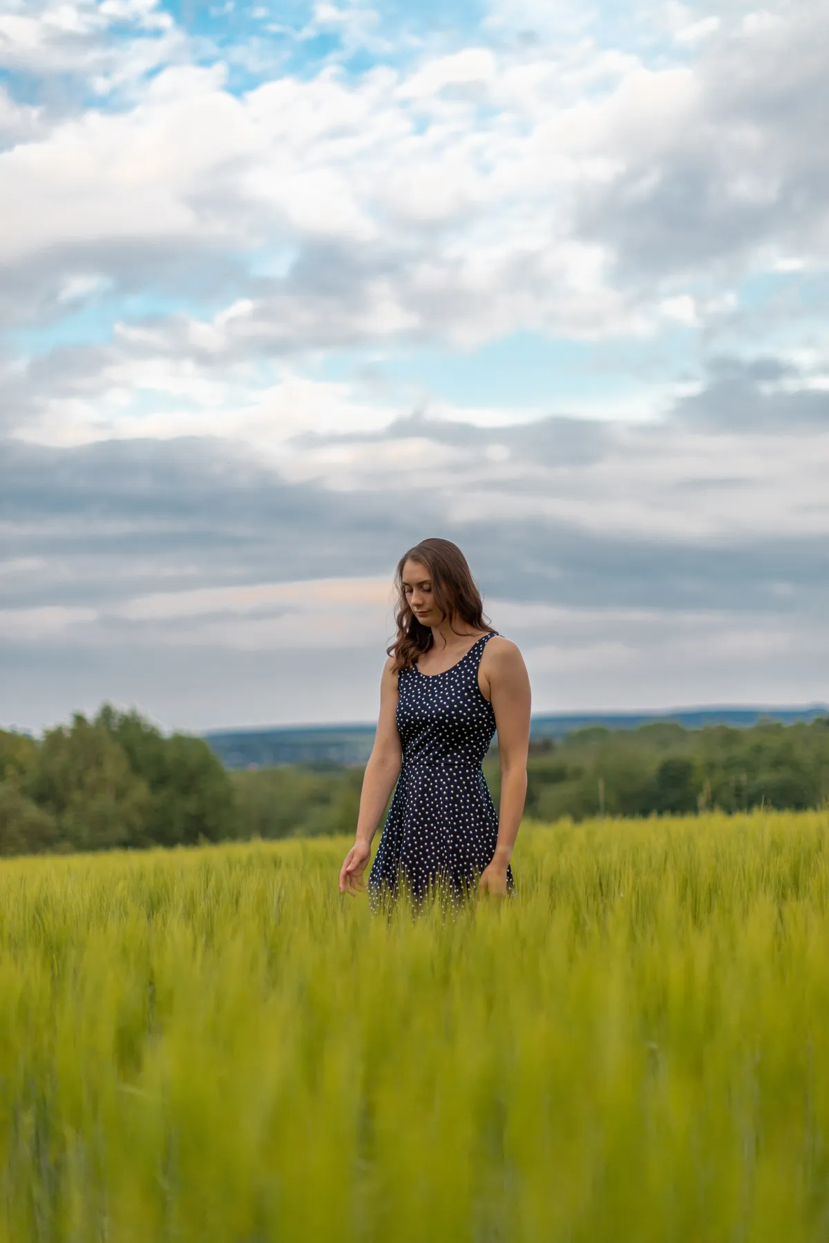 Portraitfotografie einer jungen Frau im Sommerkleid die in einem unreifen Kornfeld steht und nachdenklich nach unten schaut.