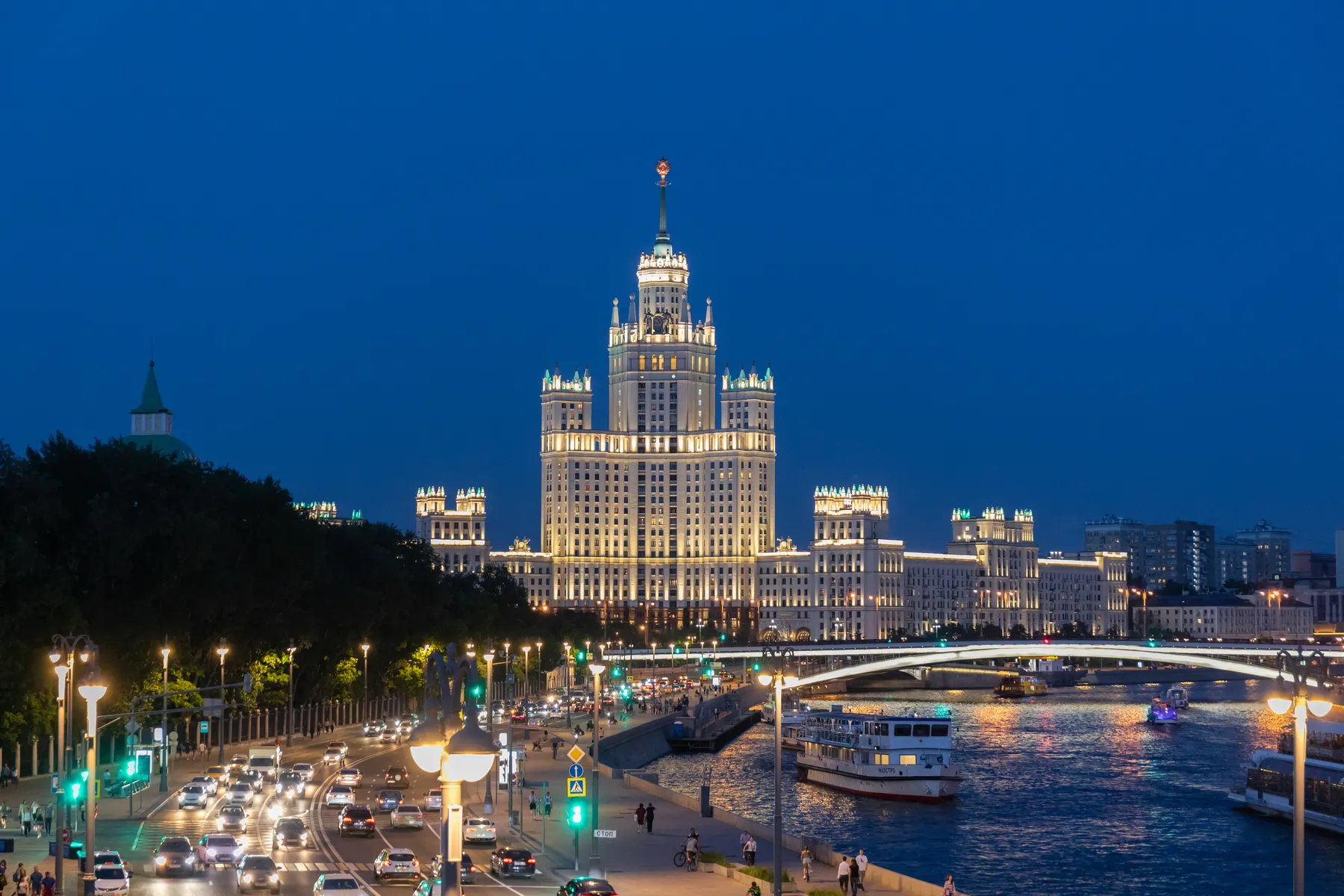 Ein hell erleuchteter, hoher stalinistischer Wolkenkratzer steht in der Abenddämmerung an der Moskva, mit einer belebten Straße und Autos im Vordergrund. Entlang des Flusses liegen Boote vor Anker, und der Himmel ist tiefblau.