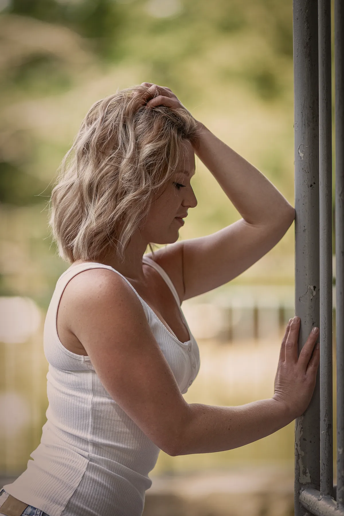 Portrait einer blonden Frau im weißen Top, seitlich stehend, Hand im Haar, nachdenklich, an Metallpfosten gelehnt, weicher Hintergrund, natürliches Licht, ruhige Stimmung, draußen, unscharfe Naturkulisse, sommerlich, intim, authentisch, ruhig, Moment