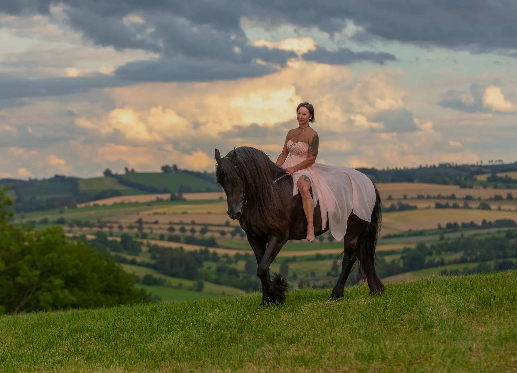 Eine Frau reitet barfuß auf einem schwarzen Pferd über eine grüne Anhöhe, umgeben von sanften Feldern und dramatischem Himmel. Fließendes Kleid, ruhige Bewegung und warmes Abendlicht vermitteln Eleganz und Freiheit, ideal für emotionale Pferde- und Hundefotografie in natürlicher Landschaft, mit zeitloser Stimmung, Tiefe, Harmonie und Ruhe für authentische emotionale Bildwelten.