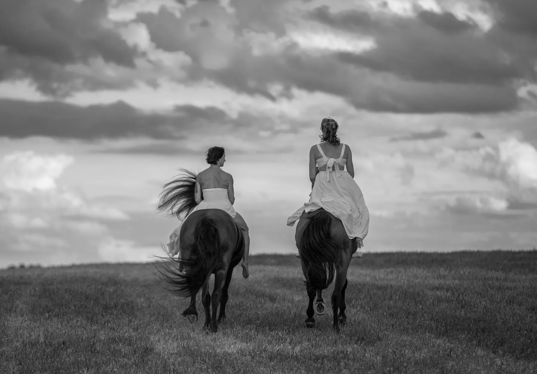 Zwei Reiterinnen von hinten bekleidet mit eleganten Kleidern galoppieren auf den Horizont zu