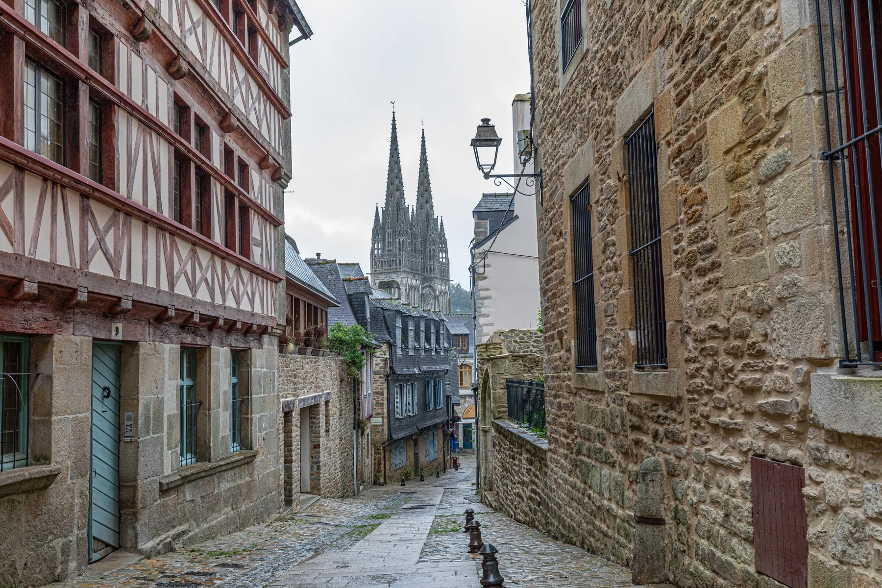 Eine schmale Kopfsteinpflasterstraße in Quimper, gesäumt von alten Stein- und Fachwerkhäusern, führt zu einer entfernten gotischen Kathedrale mit Zwillingstürmen unter einem wolkenverhangenen Himmel.